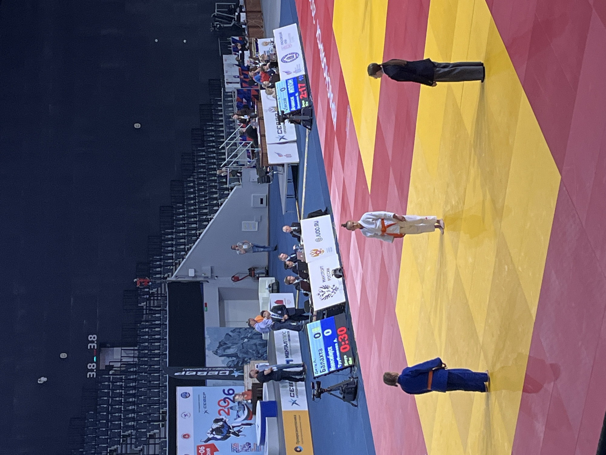 This image depicts an indoor judo competition in Krasnoyarsk, Russia. The scene is dominated by a multi-colored judo mat, composed of alternating red and yellow squares, where two female competitors and one official are standing. The competitor on the left wears a blue judogi, while the one on the right wears a white judogi with an orange belt. A female official or referee in a dark suit stands to the far right on the mat.
In the mid-ground, to the left of the mat, a digital scoreboard is visible, displaying "Балахта 0" and "Новосибирск 0", along with "Трубникова А." and a countdown timer of "0:30". To its right, an official video camera on a tripod is positioned. Further right, along the edge of the mat, several individuals in suits and casual attire are seated at long tables, acting as judges or event staff. Monitors and paperwork are visible on these tables. Another digital scoreboard in the background shows a timer of "2:17".
Various banners and signs are displayed throughout the arena. A prominent banner on the far left shows "КРАСНОЯРСК", "2026", and "СЕВЕР". Other banners feature Russian text such as "МЕНТАЛЛЮКС", "МИНОБРОНАУКИ РОССИИ", and "JUDO.RU". The background consists of dark, largely unoccupied stadium seating that ascends into a high, dark ceiling area with visible overhead lighting fixtures. Several individuals are sparsely distributed in the background, some walking or standing near the entrances.