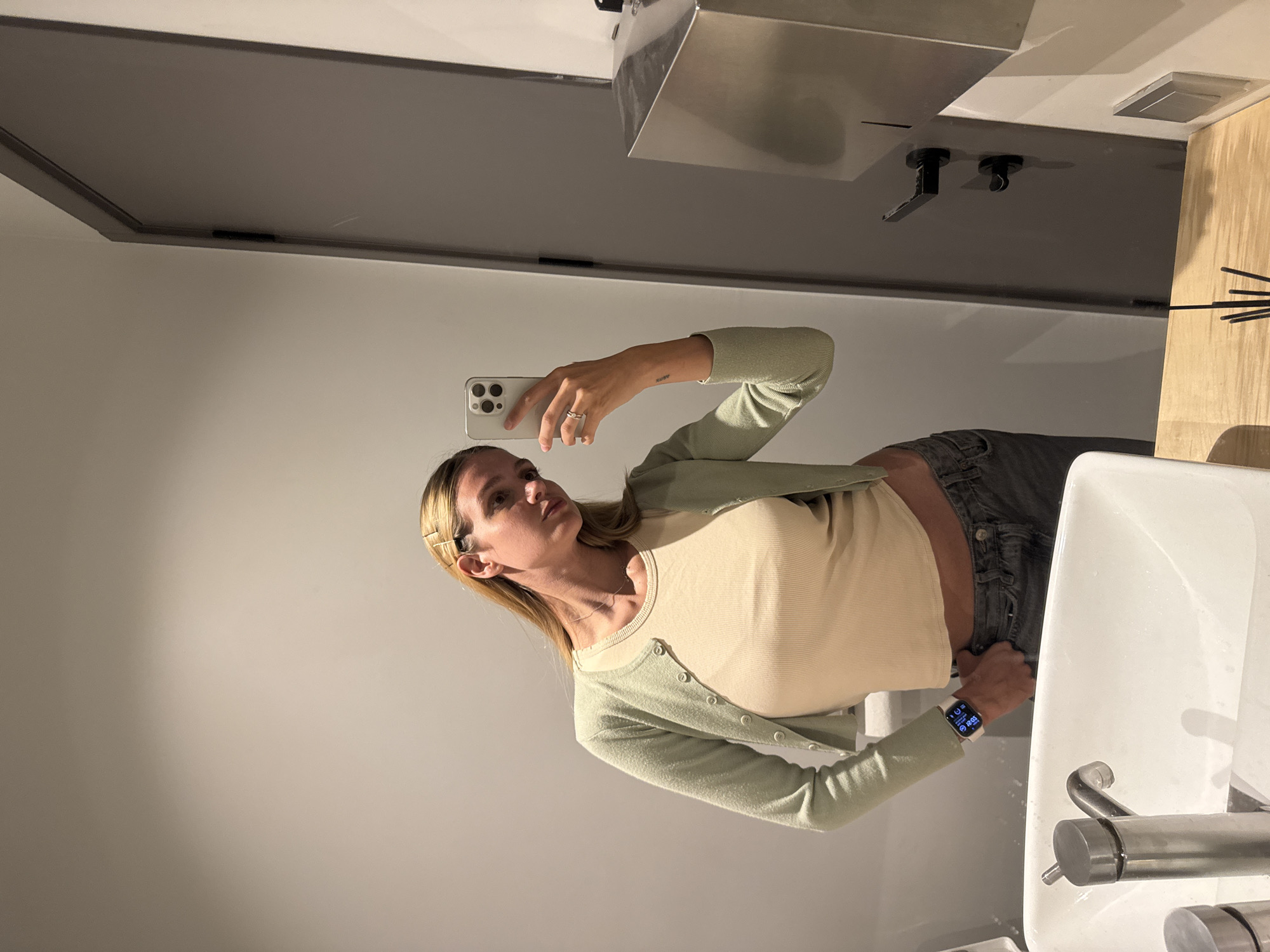 A woman takes a selfie in a bathroom. She wears a light green cardigan over a cream-colored shirt and dark grey pants. Her blonde hair is partially pulled back with bobby pins. She holds a smartphone in her right hand, taking the photo. She wears a watch on her left wrist. The bathroom features a modern sink and faucet, a wall-mounted soap dispenser, and a light-colored wood countertop. The walls are light grey. While it is impossible to determine the exact location in Tbilisi without further information, the bathroom appears to be in a modern, well-maintained building.