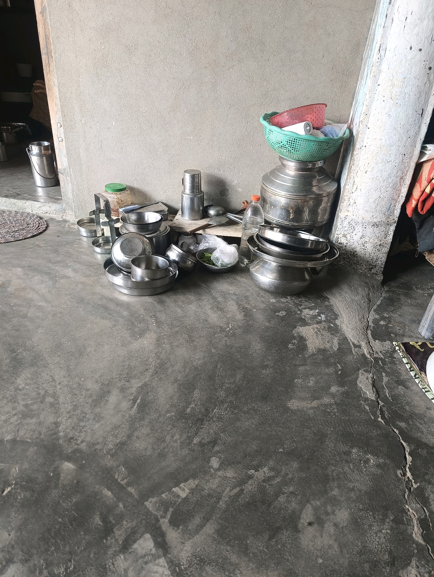 The image depicts an outdoor scene in Chhapri Bari, India, showing a collection of metal kitchenware scattered on a concrete floor. The items include pots, bowls, and a large metal container, some of which are stacked. A green plastic basket containing red plastic bowls and white cylindrical objects sits atop a large metal drum. A water bottle with an orange cap is also visible. A rough concrete wall forms the backdrop, with a white-painted pillar to the right. The floor shows signs of wear and a crack. The environment appears to be an unkempt outdoor area, possibly a kitchen or storage space. No people are present in the frame.