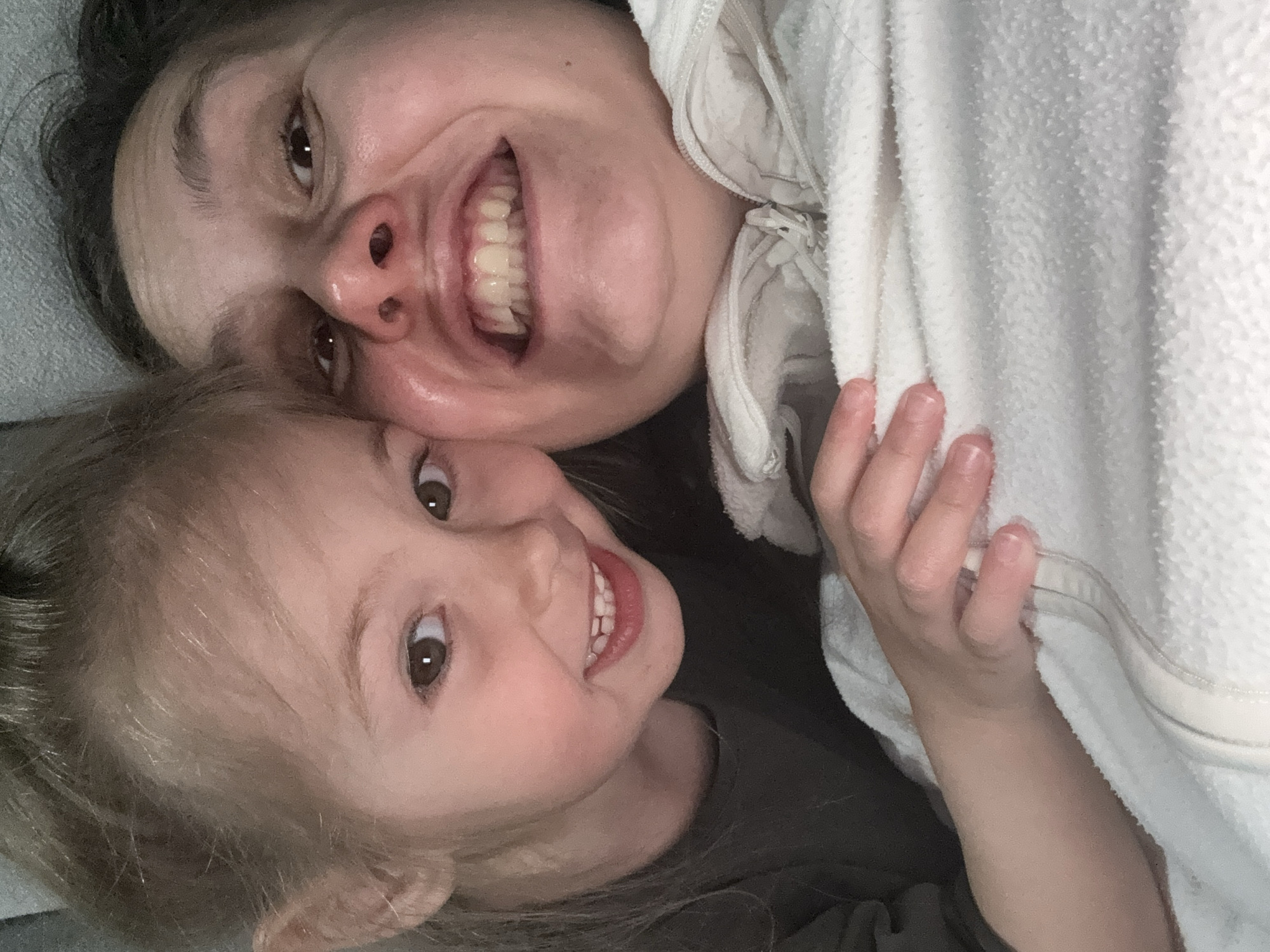 A selfie shows a woman and a child, likely a mother and daughter, lying down and smiling at the camera. The woman has dark hair and is partially covered by a white, possibly hooded, garment. The child, who is positioned closer to the bottom of the frame, has light hair and is wearing a dark-colored shirt. The background appears to be a plain, light-colored surface. The image was not taken in Zelenogradsk, Russia.