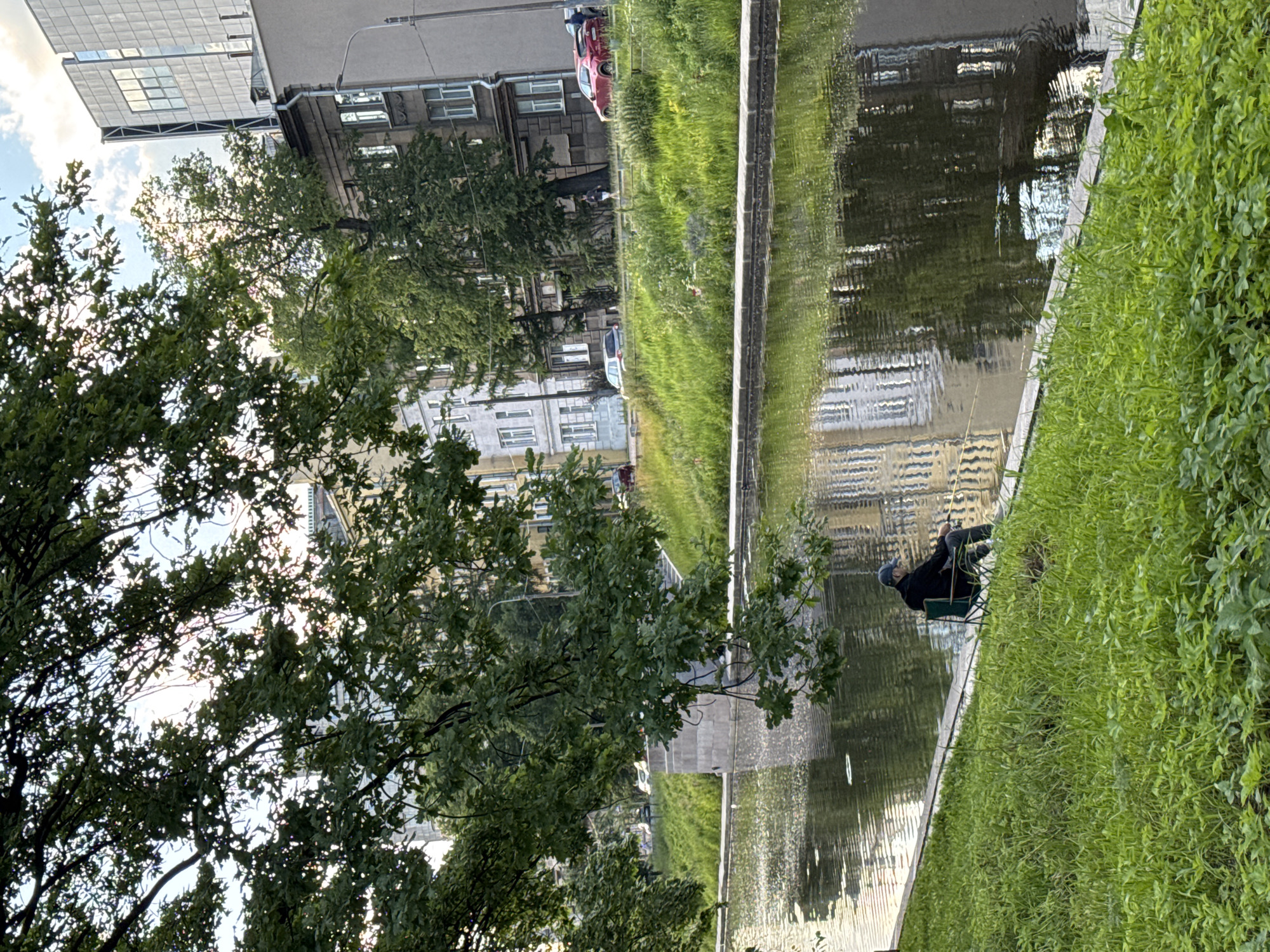 The image shows a man fishing on the bank of a canal in Saint Petersburg, Russia. The man is sitting in a green folding chair and holding a fishing rod. The canal is surrounded by lush green grass and trees. In the background, there are buildings of various architectural styles, some modern and some older. The water in the canal reflects the sky and the surrounding buildings. The overall impression is one of tranquility and peacefulness in an urban setting.