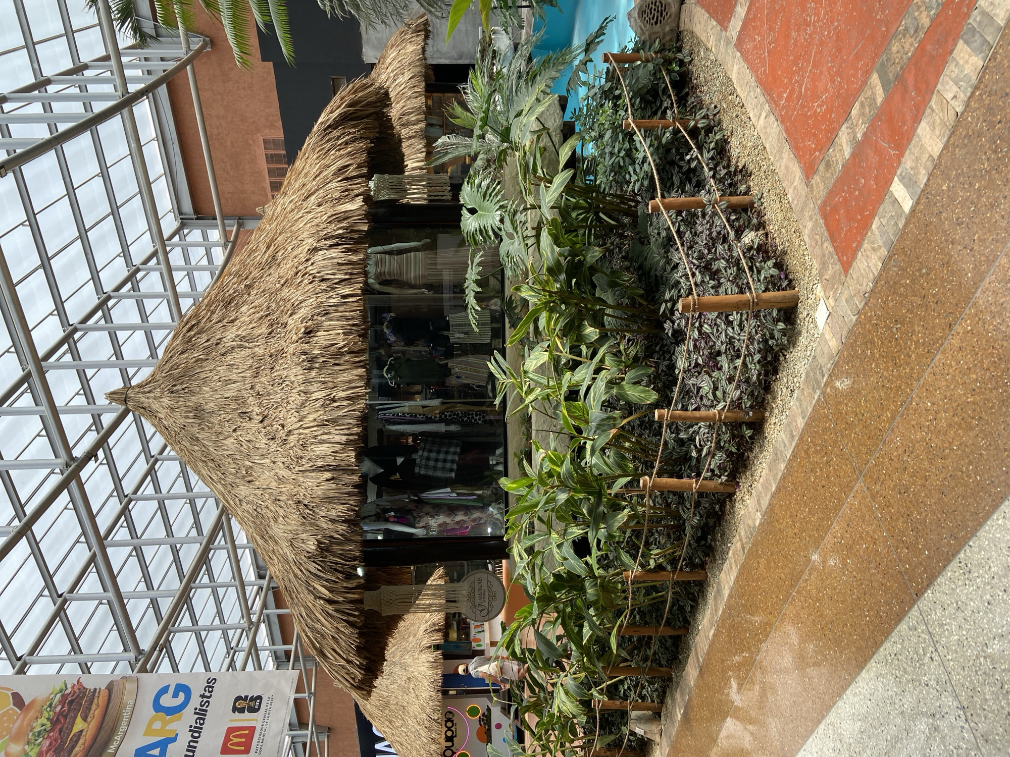 This image depicts an indoor shopping mall with a tropical-themed display. The focal point is a store front designed to resemble a thatched-roof hut, complete with lush greenery and a small water feature that appears to be a pool. The hut has large glass windows displaying mannequins dressed in clothing, suggesting it is a boutique.
To the left of the main hut, a smaller, similar structure is visible. The surrounding area features a pathway made of polished aggregate concrete in shades of brown and orange. The display is cordoned off with wooden posts and rope, creating a clear boundary.
In the background, a large banner with the text "McArgentina" and "ARG Mundialistas" is displayed, along with the McDonald's logo and a FIFA emblem, indicating a promotion related to the World Cup and sponsored by McDonald's. The ceiling of the mall is a complex metal and glass structure, allowing natural light to filter in.
A person is visible walking in the distance near the smaller hut, dressed in casual attire. The overall environment suggests a commercial space designed to evoke a resort-like atmosphere within a mall in Pampatar, Venezuela.