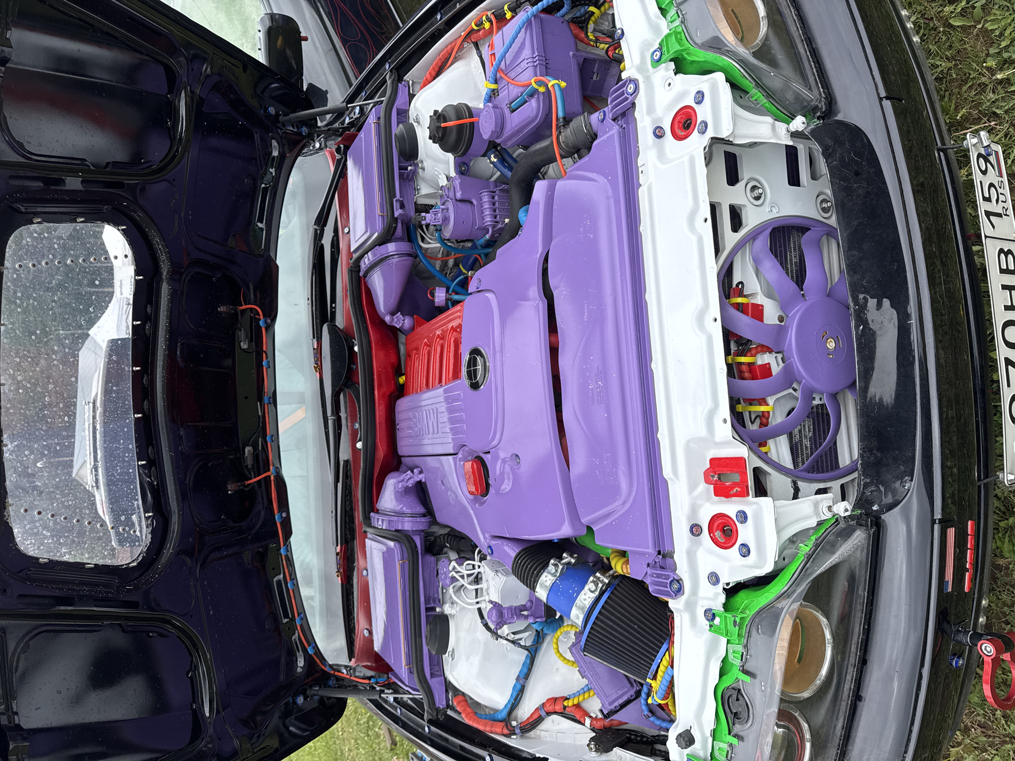 The image shows the engine bay of a car in Chaikovsky, Russia. The engine and surrounding components are painted in various colors including purple, white, red, and green. A Russian license plate is visible.