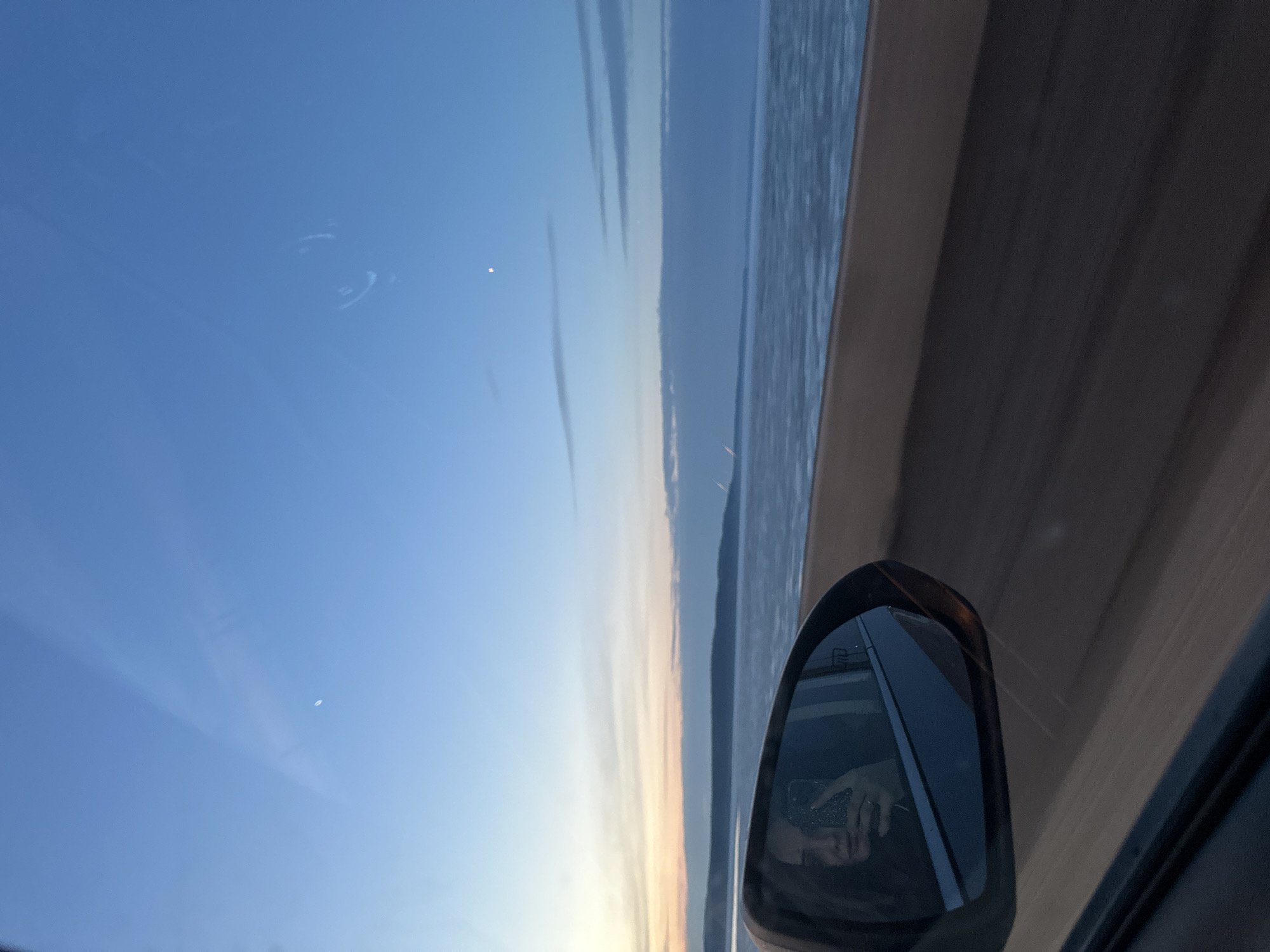 The image shows a view from inside a moving vehicle. The sky is visible through the windshield, transitioning from blue to a light orange/yellow near the horizon, suggesting either sunrise or sunset. There are faint cloud formations. The side view mirror of the vehicle reflects a person holding a phone, presumably taking the photo. A glimpse of water (possibly a river or lake) is seen through the side window. The location might be near the Kama River, as it is the primary water feature in Chaikovsky, Russia, based on the orientation of the sky and the possible waterbody. The interior of the car is dark, but the window frame and parts of the dashboard are visible.