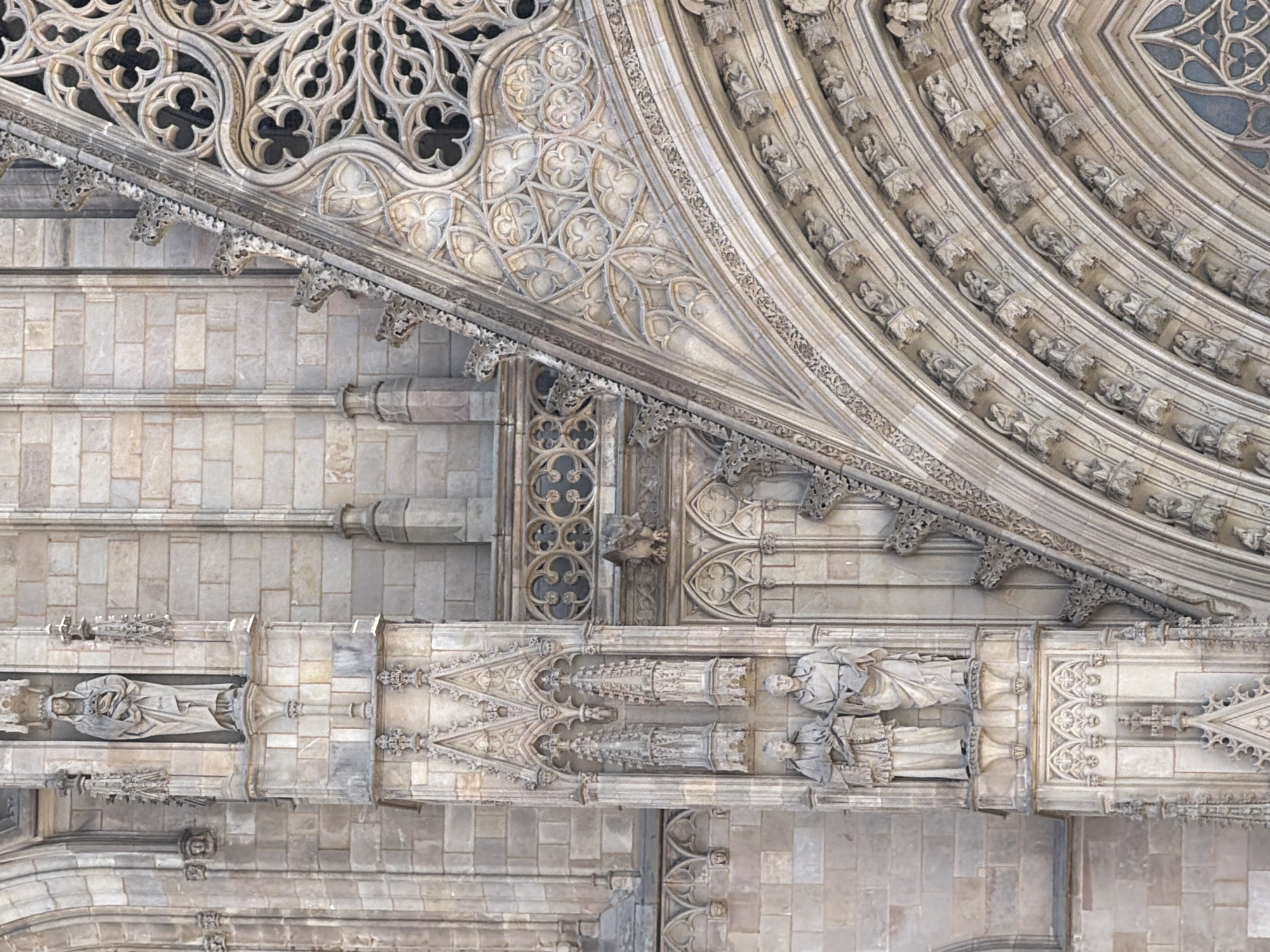 The image shows a close-up, eye-level view of the exterior facade of the Barcelona Cathedral (Catedral de Barcelona) in Barcelona, Spain. The architecture is Gothic, characterized by intricate stone carvings and detailed ornamentation. The focus is on the upper sections of the cathedral, specifically the ornate stonework of a large arched window and the vertical elements flanking it.
On the left side of the image, a niche contains a statue of a robed figure holding an object in its hands. Below this, on a projecting stone pedestal, are two more statues of robed male figures, one holding a scroll or book. These statues are positioned within deeply recessed architectural niches adorned with Gothic tracery.
The central and right portions of the image are dominated by a large, elaborate rose window or tracery window, with intricate geometric patterns resembling quatrefoils and trefoils. The stone surrounding the window is carved with curvilinear designs and small architectural motifs. Below the window, a series of arched recesses are visible, each containing a small carved figure, possibly angels or saints, appearing to be in various poses. The overall impression is one of immense detail, craftsmanship, and the weathered patina of ancient stone. The lighting suggests it is daytime, with shadows highlighting the depth of the carvings.