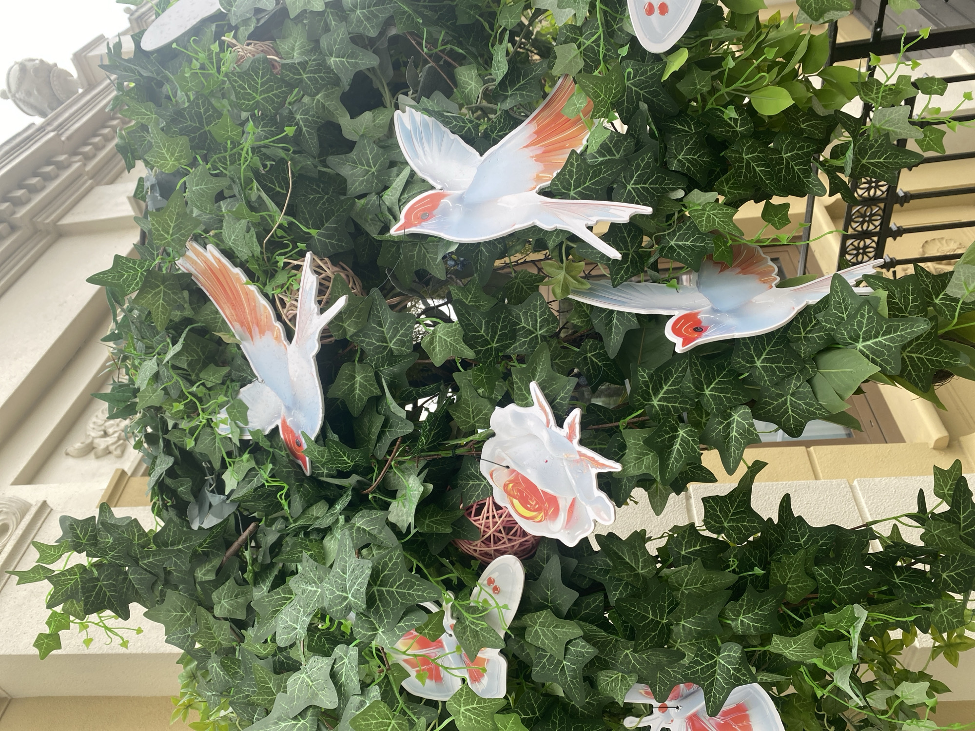 The image depicts an artificial ivy vine adorned with several decorative cutouts of birds and flowers. The birds are depicted in flight, with white bodies and orange accents on their wings and tails. One bird in the center right of the image is facing upwards with its wings spread, while another is on the top right with its wings tucked in. Below and to the left, another bird is in a similar pose to the center right one. There are also some flower-shaped cutouts, one with red and yellow detailing, and another more abstract white and red shape. A small woven pink sphere is visible amongst the foliage. The background shows part of a building with creamy-colored walls and decorative architectural elements. The scene appears to be outdoors, possibly a balcony or an entranceway, with a black metal railing visible at the bottom. The image is taken from a slightly high angle, looking down into the foliage. The lighting suggests an overcast day. There are no people visible in the image. The location is identified as Khimki, Russia.