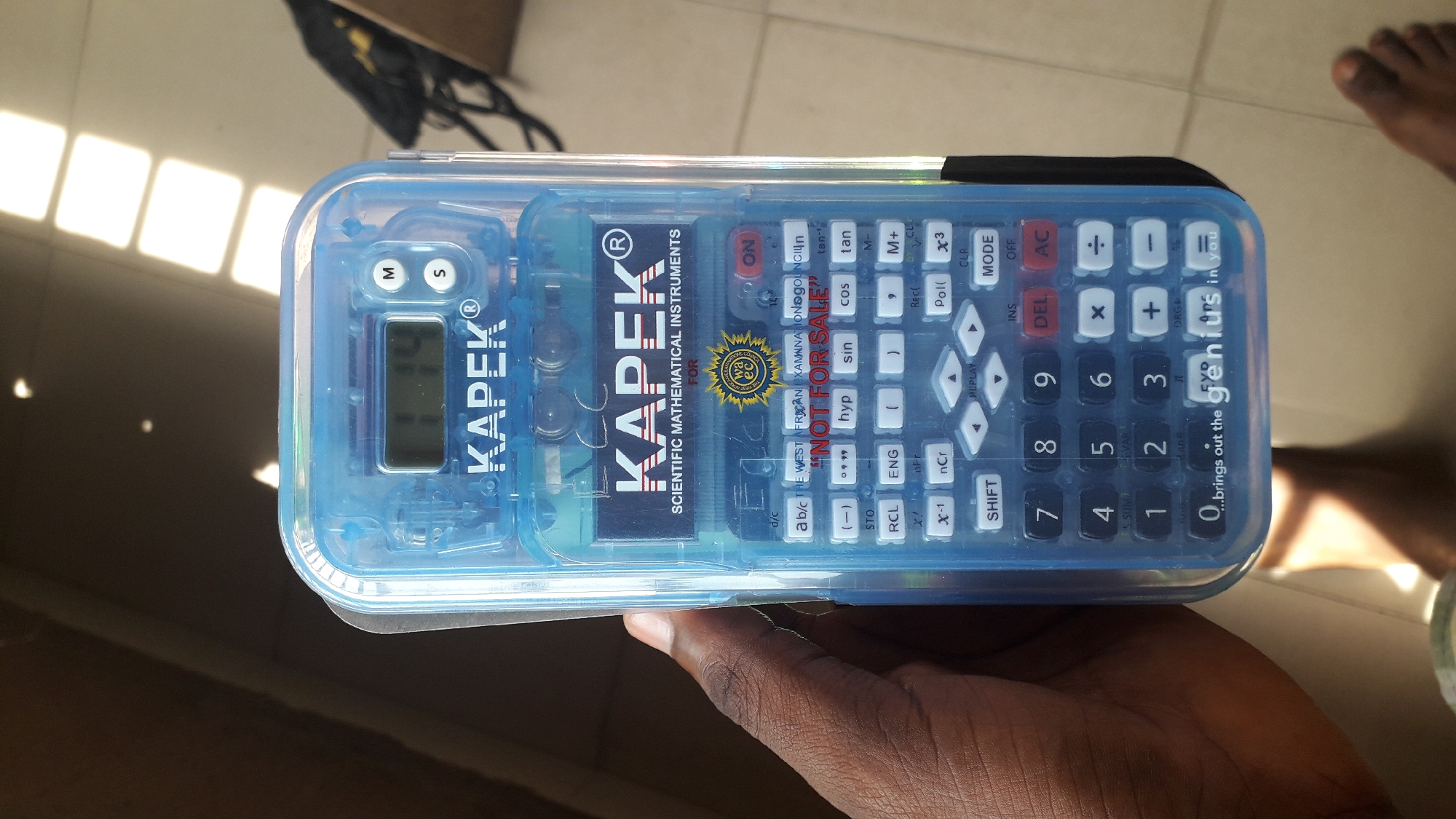A close-up photograph depicts a hand holding a KAPEK® scientific calculator in a clear blue plastic case. The calculator is resting in the palm of a dark-skinned hand. The calculator features a display screen showing a number. The calculator functions read “KAPEK Scientific Mathematical Instruments for”. “The West African Examination Gouncin” and the phrase "NOT FOR SALE" are printed. Additional function buttons visible include trigonometric functions, logarithms, and other scientific notation, as well as basic mathematical operators. The product has the tagline, "...brings out the genius in you". The calculator’s ON button is red. The image includes a glimpse of someone’s feet in the top right corner and a black bag and brown box is out of focus in the top left corner. The photograph appears to have been taken in an interior space with light reflecting on the floor. There is no explicit indication of its location in Nkpor, Nigeria.
