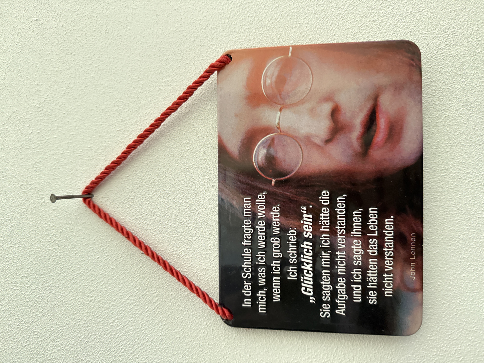 A plaque with a quote by John Lennon hangs on a textured, off-white wall, suspended by a red braided cord attached to a nail. The plaque features a close-up portrait of John Lennon wearing round glasses. The quote, written in white text on a black background, reads: "In der Schule fragte man mich, was ich werde wolle, wenn ich groß werde. Ich schrieb: 'Glücklich sein'. Sie sagten mir, ich hätte die Aufgabe nicht verstanden, und ich sagte ihnen, sie hätten das Leben nicht verstanden. John Lennon". The plaque is positioned at eye level. The environment is indoor, likely a home or a public space.