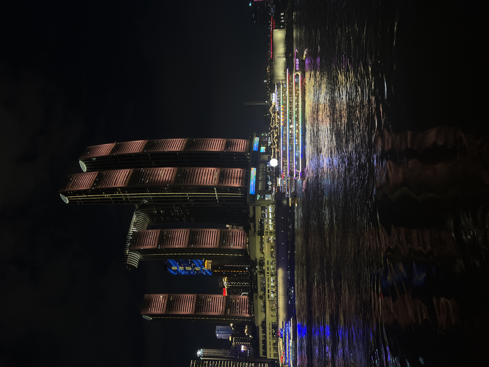 This nighttime photograph captures a waterfront view of the Nan'an District in Chongqing, China. The scene is dominated by a cluster of illuminated skyscrapers, featuring a prominent architectural design with a horizontal connecting structure between two of the tallest buildings. These buildings are lit with warm, reddish-orange and yellow lights, highlighting their verticality and intricate facades. In the foreground, a wide river reflects the vibrant lights from the cityscape and two illuminated passenger boats. The boat on the left is primarily lit with blue and white lights, while the larger boat on the right displays a colorful spectrum of lights along its decks. The water is choppy, creating a dynamic surface that diffuses the reflections. The sky is dark, indicating the late hour, with minimal cloud cover. Several other buildings and a bridge with red lights are visible in the background, contributing to the overall urban nightscape. There are no visible people in the frame.