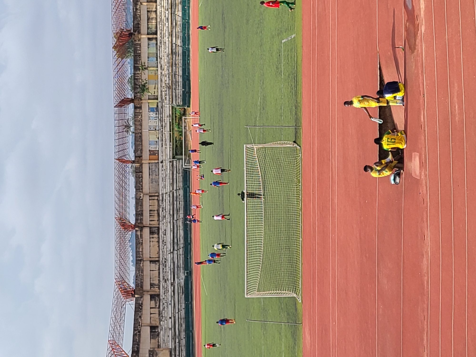 The image shows an outdoor sports stadium with a soccer field and a running track, a stadium building on the left and cloudy sky above. On the soccer field, multiple players in red and blue uniforms are engaged in a game. A soccer goal is visible. Three people dressed in yellow uniforms are sitting on the running track in the right. They are looking toward the soccer field.
