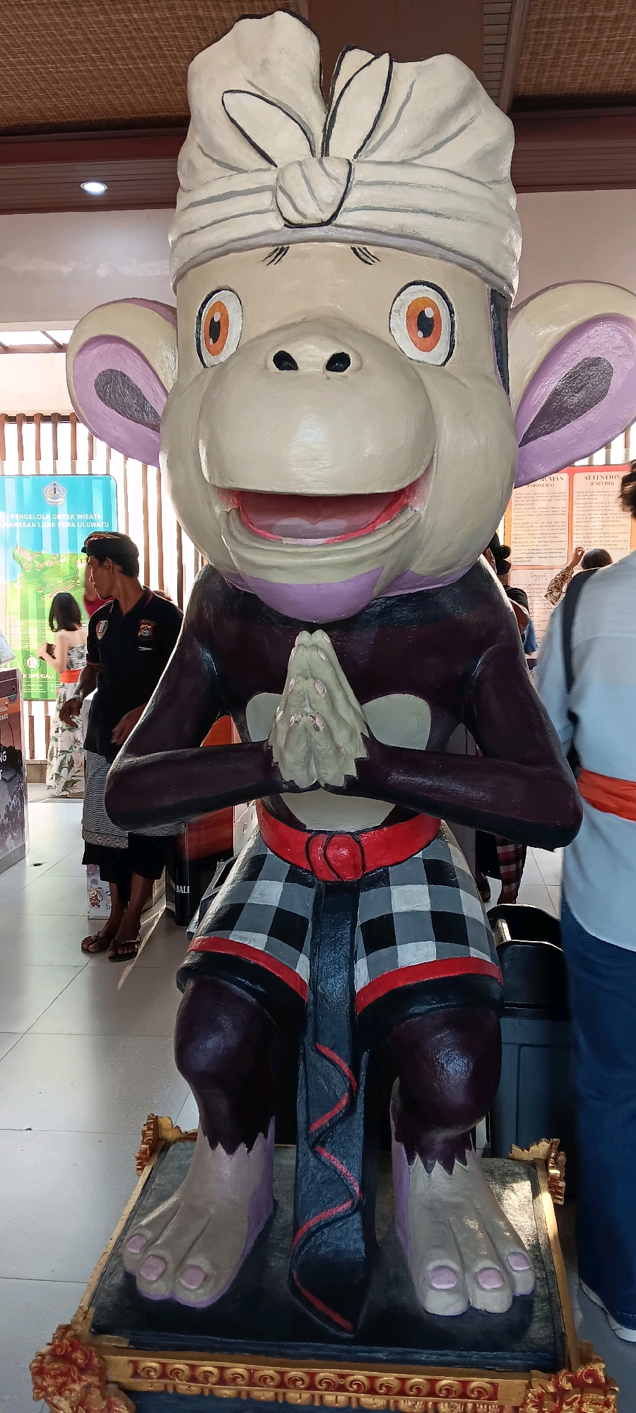 The image shows a full-body statue of a monkey wearing a traditional Balinese checkered sarong, a red belt, and a white head covering. The monkey has large, expressive eyes and hands clasped in front of its chest in a greeting gesture. The statue is placed on a decorative golden base. In the background, there are people, informational posters, and a wooden ceiling structure. The scene appears to be indoors, possibly at a tourist attraction or cultural site in Kuta Selatan, Indonesia, given the attire and the informational signage.