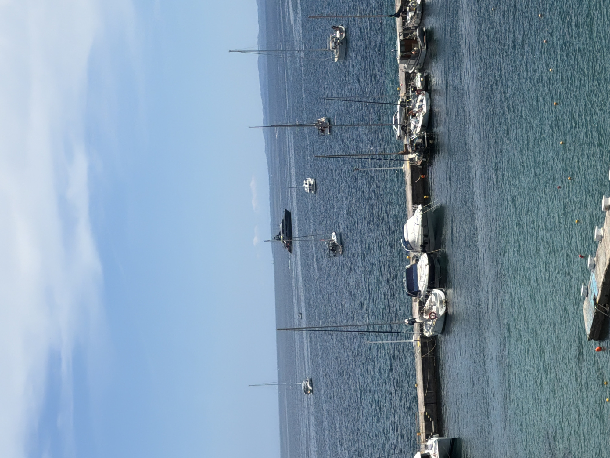 The image, when viewed in its correct landscape orientation, presents a tranquil harbor or marina scene, likely in Corfu, Greece. The setting is dominated by a clear, bright blue sky with faint, wispy white clouds visible on the left side.
A calm, blue-green body of water fills the majority of the frame. On the right side, a long, concrete pier extends horizontally, serving as a mooring point for numerous boats. Several sailboats and a few smaller motorboats are neatly docked along this pier. Their tall masts stand upright, and some details like lifebuoys are discernible on their decks. The water near the pier appears lighter and clearer.
Further out in the deeper, darker blue waters on the left, several other sailboats and what appears to be a larger, dark-hulled vessel (possibly a yacht) are anchored or moored. These boats are scattered across the water, giving a sense of depth to the bay or sea. A distant, hazy coastline or island stretches across the horizon in the mid-ground, providing a backdrop to the boats.
Small yellow and orange spherical buoys are visible floating in the water, likely indicating mooring spots or navigation markers. The water surface shows only gentle ripples, suggesting very calm weather conditions with little to no wind.
The scene depicts a peaceful daytime environment, most likely late morning or early afternoon given the bright sunlight. There are no clear, identifiable individuals, although small figures might be present on some of the docked boats. No discernible text is visible within the image. The overall impression is one of serene maritime leisure.