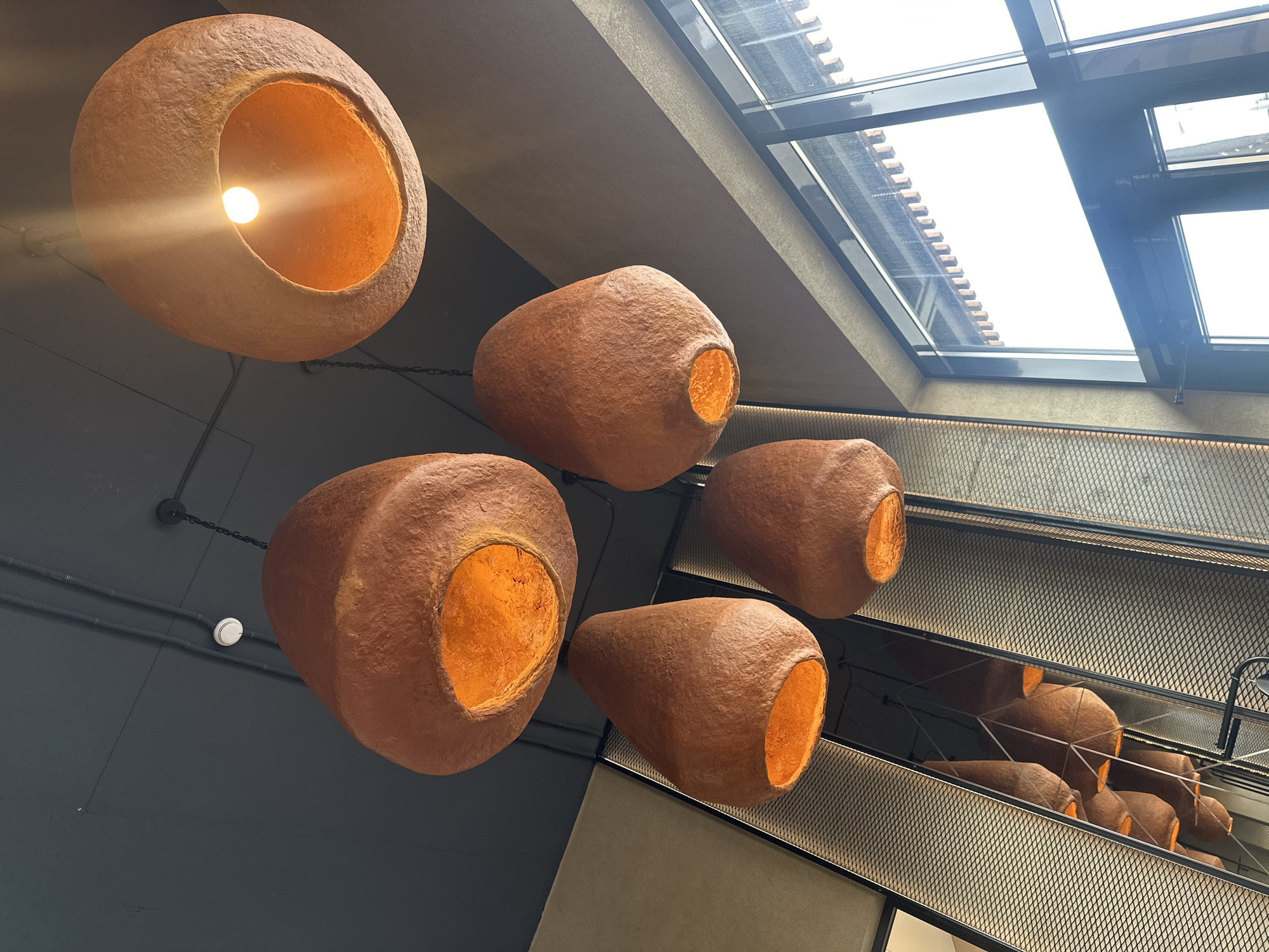 An interior shot depicts a modern commercial or public space in Усады, Russia, characterized by unique architectural lighting and design elements. Six large, organic-shaped pendant lights with textured, reddish-brown exteriors hang from a dark grey ceiling. The individual light fixtures vary in shape, from ovoid to teardrop, and emit a warm, amber-toned light from their exposed incandescent bulbs or LED filaments, which illuminate their internal surfaces in a vibrant orange hue.
The dark grey ceiling features visible black electrical conduits and a small, circular white smoke detector. Chains suspend the pendant lights from the ceiling. To the left, a textured wall, possibly constructed from metal mesh, stands adjacent to a tall, narrow mirrored panel that reflects additional pendant lights, creating an illusion of depth and more fixtures. The right side of the image is dominated by large, dark-framed windows that allow bright, undifferentiated natural light into the space. A light-colored, possibly beige or light grey, wall section is visible above the windows, adding to the varied material palette. No individuals are present in the scene.