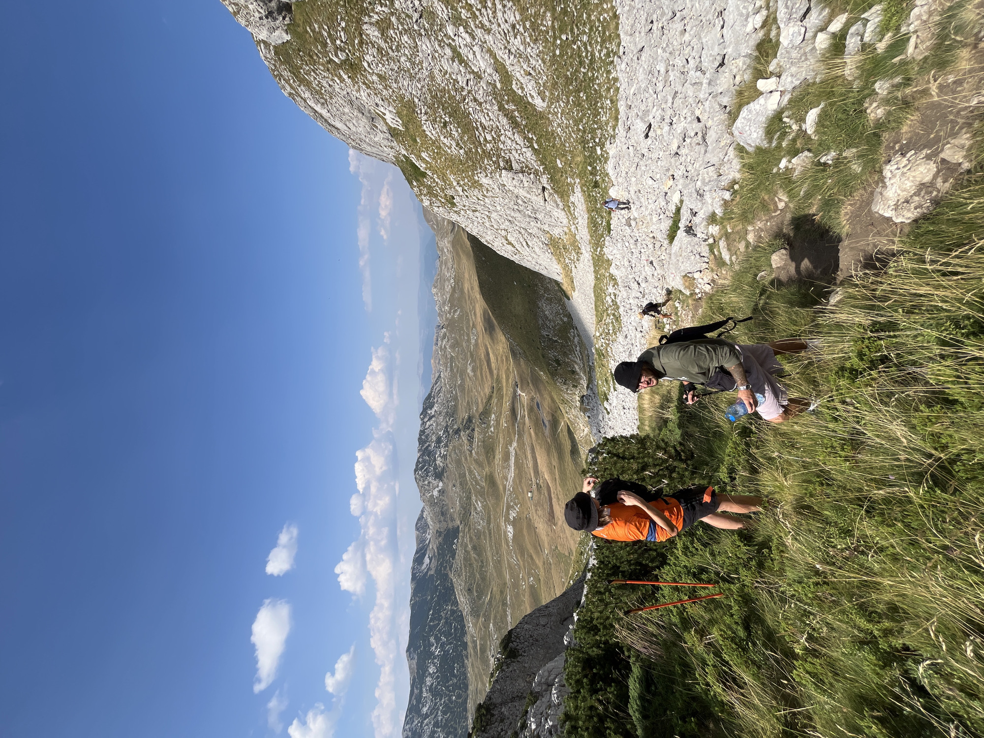 This is a vertical, eye-level, outdoor, medium-shot photograph of a mountainous landscape in Boricje, Montenegro. The sky is a vibrant blue with scattered white clouds. In the foreground, two hikers stand in tall, green grass. The hiker on the left wears an orange shirt and black shorts. The hiker on the right wears a green hooded jacket, grey shorts, and a black beanie. They both appear to be looking down. In the mid-ground, a rocky, mountainous terrain stretches out, with sparse vegetation and a winding path visible. Several other hikers can be seen further down the path. The background features rolling hills and distant mountains under the expansive sky. The overall scene depicts a group of people engaged in hiking in a scenic, natural environment.