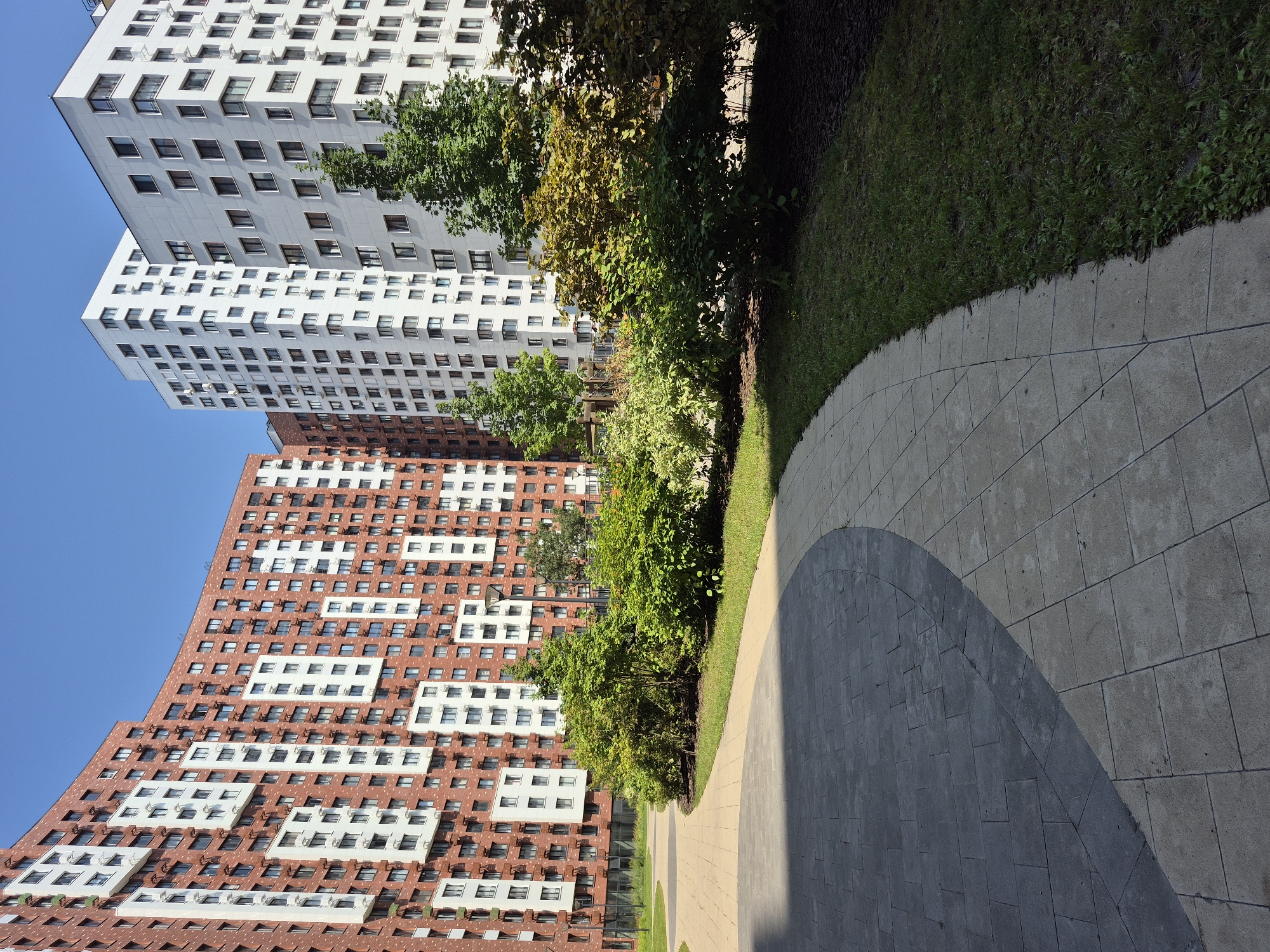 The image depicts the exterior of a modern residential complex in Moscow, Russia. The architecture features a mix of curved and straight buildings with facades of red brick and white panels. The buildings are tall, multi-story structures with numerous windows. In the foreground, a paved walkway curves through a landscaped area with green grass and various bushes and trees. The sky is clear and blue, indicating a sunny day. There are no people visible in the image. The scene appears to be a well-maintained urban environment.