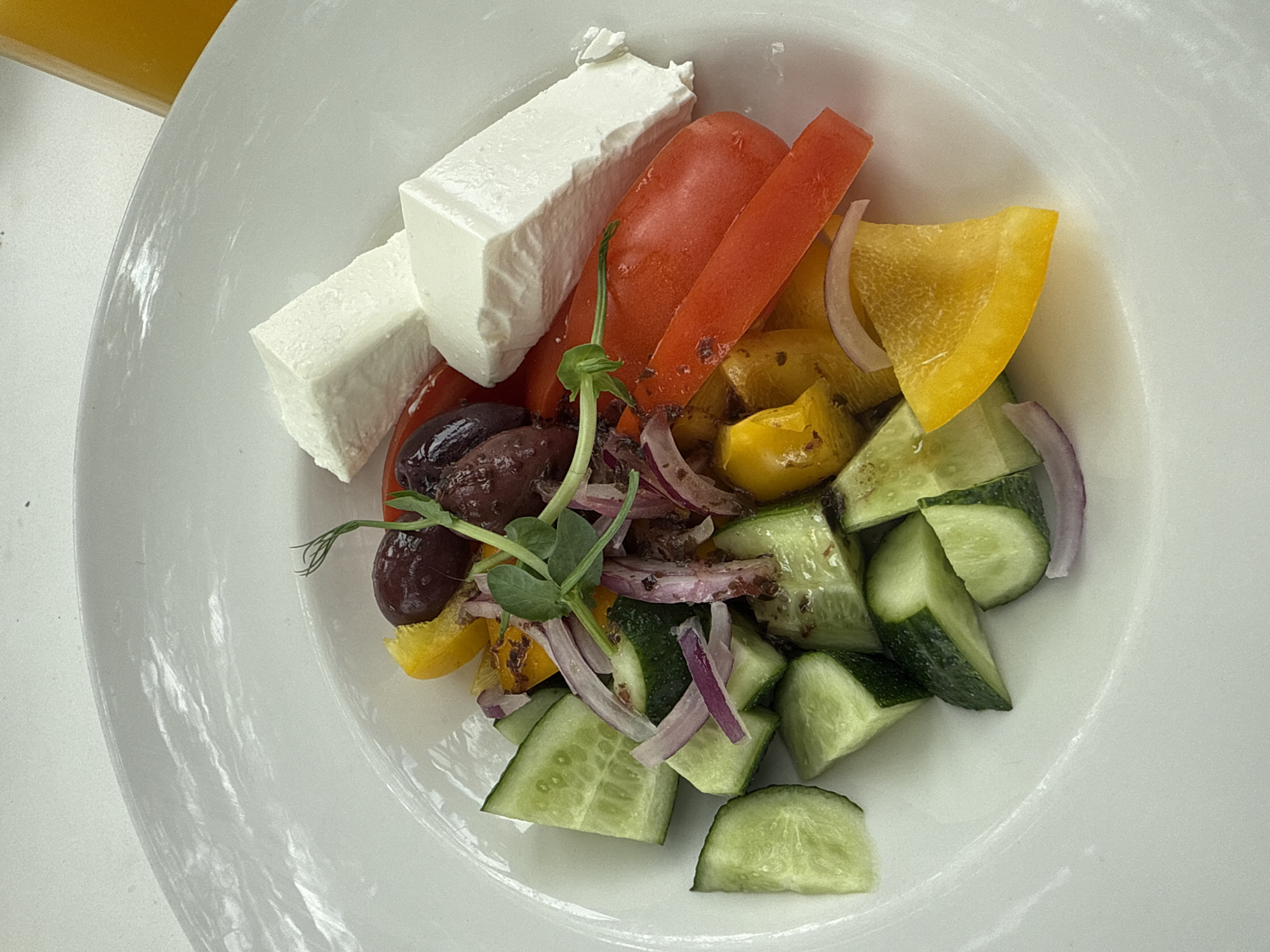 In Komarovo, Russia, a white, circular plate is presented on a white surface. The plate contains a Greek salad with feta cheese. The salad is composed of sliced cucumbers, red onions, Kalamata olives, red bell peppers, and yellow bell peppers. Two large, rectangular pieces of feta cheese are placed on top of the salad. The ingredients appear fresh and are arranged artfully on the plate. A small sprig of green herb is also visible, adding a touch of garnish. The lighting suggests an indoor setting, possibly a restaurant.