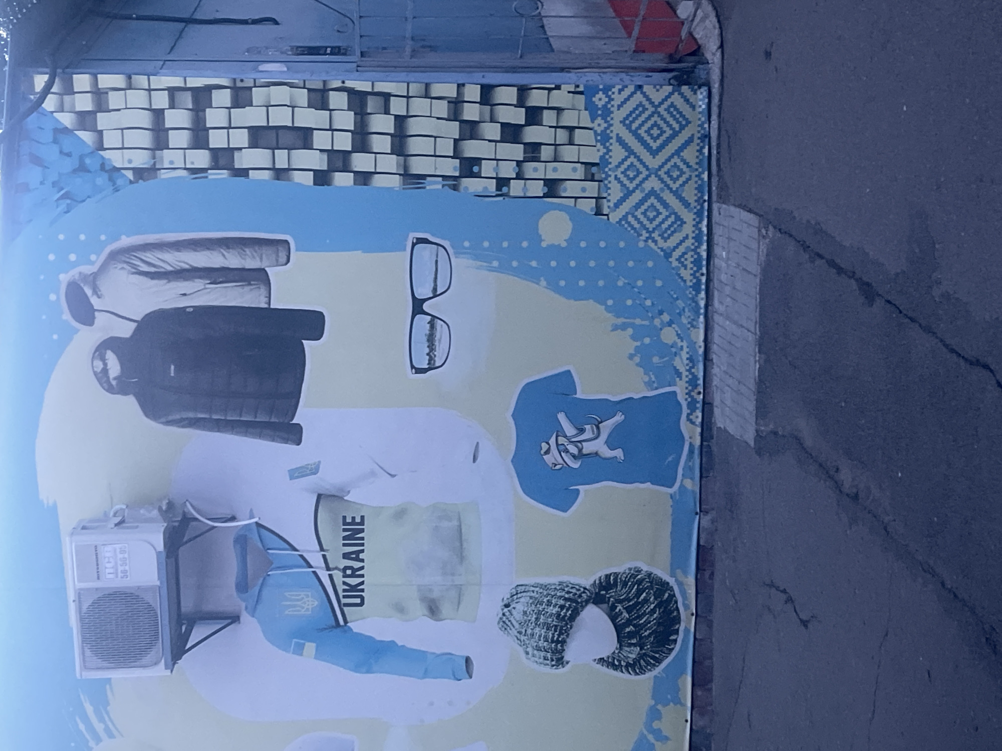 The media file is a photograph of a large mural painted on the side of a building. The mural depicts various clothing items, including jackets, a hoodie, and a t-shirt, all in shades of blue, white, and yellow. The clothing appears to be modern and casual. Some of the garments feature the Ukrainian trident emblem and the word "UKRAINE." The mural also includes graphics of a pair of glasses and a knit hat. The overall design is colorful and abstract, with a background featuring geometric patterns and a light blue wash. An outdoor air conditioning unit is mounted on the wall below the mural. The scene is outdoors, with a paved surface visible to the right of the mural, suggesting an urban or commercial setting. The lighting indicates it is daytime.