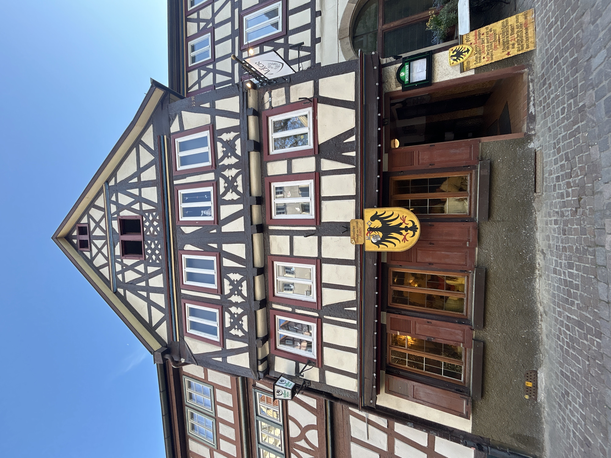 Here is a description of the image:
The image shows a close-up, angled shot of a traditional German half-timbered building, possibly a restaurant or inn, set against a clear blue sky. The building features a distinctive Fachwerk style, characterized by exposed timber framing filled with plaster or brick. The timber is dark brown, creating a striking contrast with the white infill. Windows are framed in white and red. Signboards bearing German text hang from the building, as well as signs with eagles on them. The structure appears to be located on a narrow cobblestone street. The light suggests it is daytime.