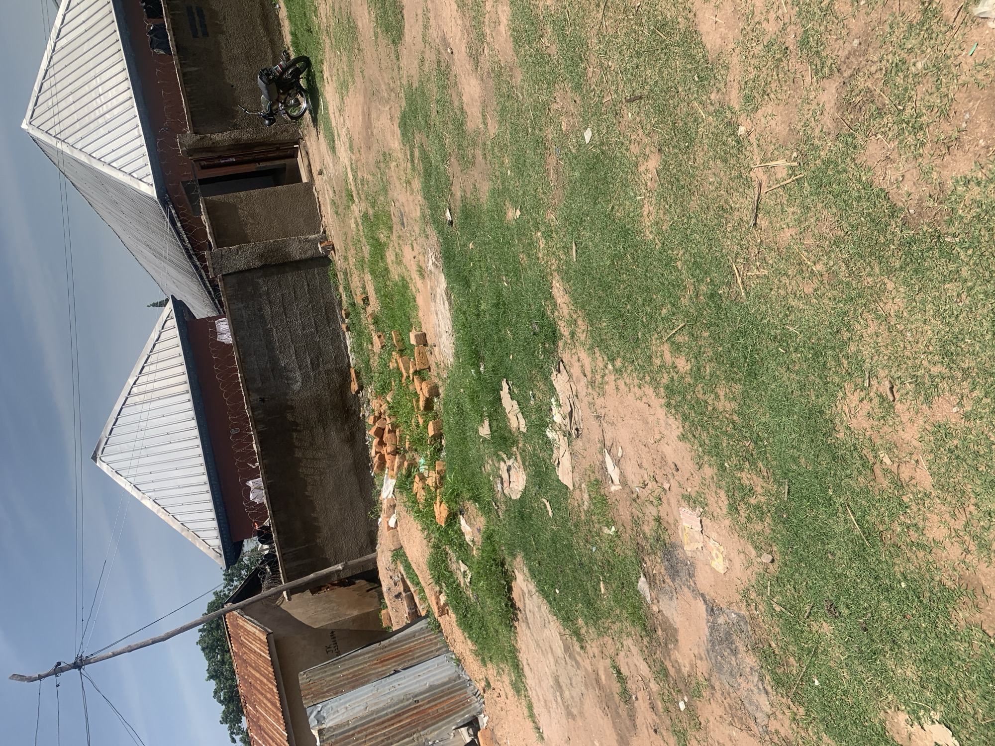 The image captures a dusty, outdoor scene in Jauro Yinu, Nigeria, under a bright, partly cloudy sky. In the background, two buildings with dark red walls and prominent, corrugated metal roofs are visible, separated by a rough, concrete wall. The wall is topped with barbed wire. To the left, a smaller, weathered building with a rusty corrugated metal roof stands next to the larger structures. A utility pole with multiple wires extends from the left side of the frame. In the foreground, a patch of sparse green grass grows on reddish-brown earth. Scattered across the ground are a pile of red bricks, some pieces of debris, and a motorcycle parked on the grass near the entrance to one of the buildings. The overall impression is one of an unpaved, possibly semi-constructed or developing area.