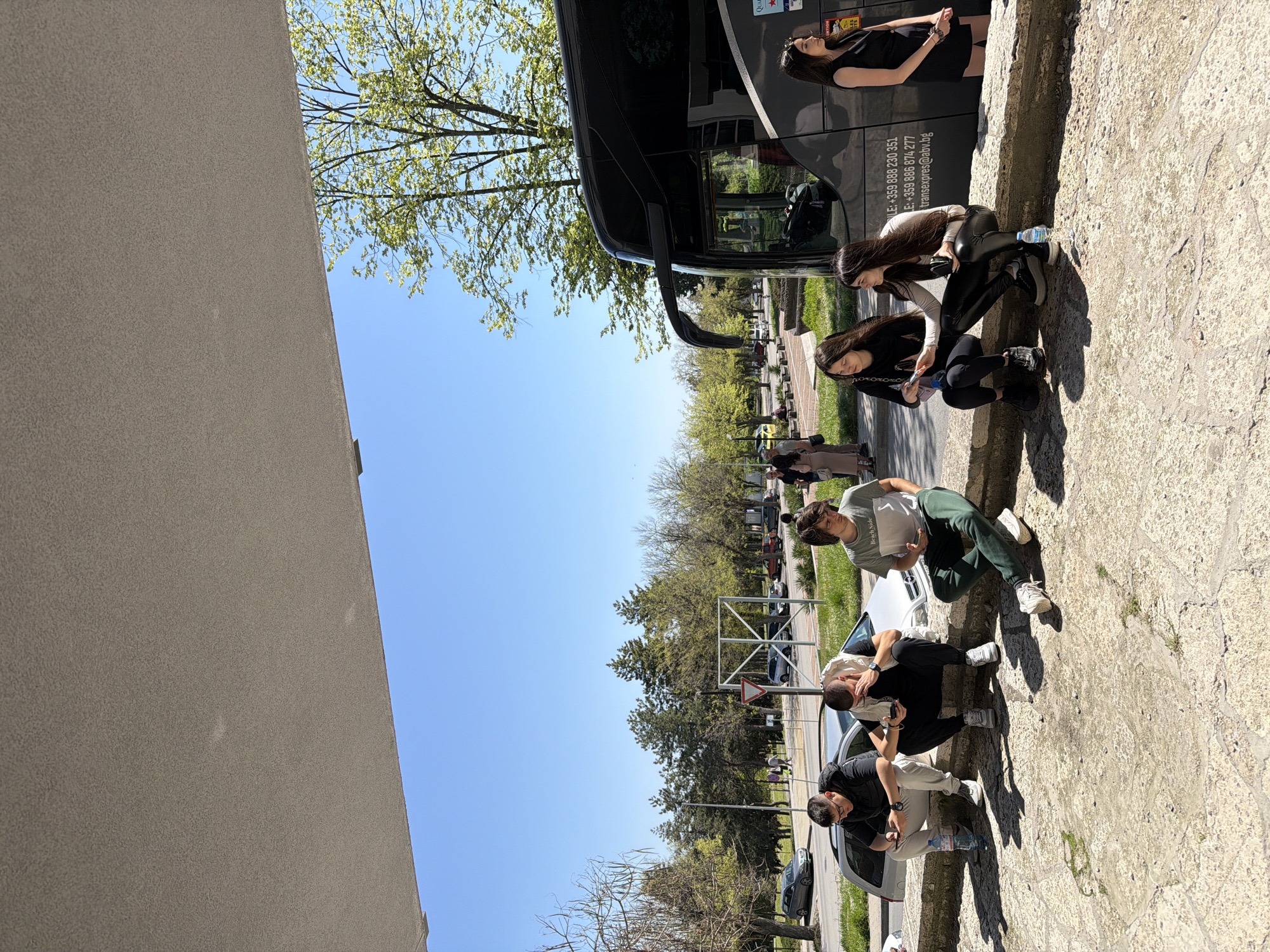 This image captures an outdoor scene on a bright, sunny day, likely in Shumen, Bulgaria, as indicated by the provided context and the Bulgarian country code in the visible text. The setting appears to be an urban or suburban street or parking area, with a building wall on one side and a clear blue sky overhead.
Several young adults are present. On the right, three women are gathered near a black bus; one stands, dressed in black, while two others crouch or sit on a low concrete wall. Further down on the left, three men are also crouching or sitting near parked cars, one wearing a grey t-shirt with "Beach Side" printed on it, and another in a black top. Most individuals are casually dressed and appear to be looking at their phones or casually interacting, suggesting a period of waiting or a break.
Notable objects include a large black bus, several cars (including a white one), a textured beige building wall, and trees with fresh green leaves, indicating spring or early summer. A small water bottle is visible next to one of the men.
Visible text on the side of the bus includes contact information: "TLE: +359 888 230 351", "E: +359 886 874 277", and "transexpres@abv.bg". Partial text "QUA" is also seen on the bus, and "Beach Side" is on a man's t-shirt. The clear sky and strong sunlight suggest it is daytime with pleasant weather.