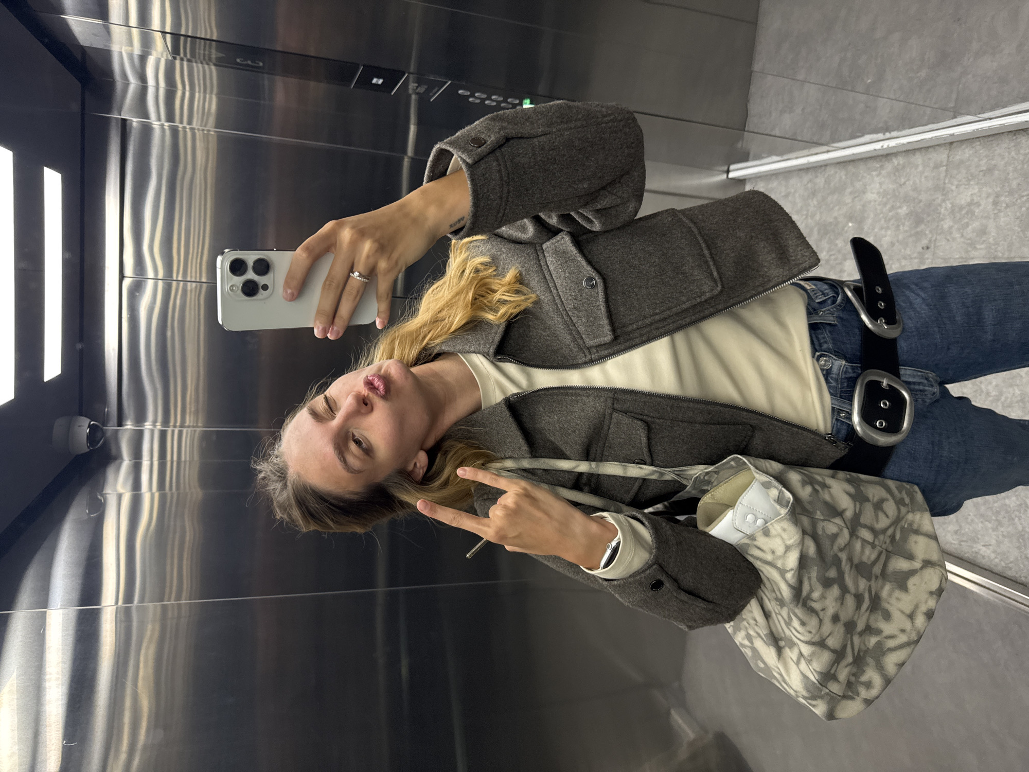 A young woman is taking a selfie in an elevator. The elevator interior is stainless steel. She is holding a white smartphone in her right hand, the back camera visible. She has blonde hair and is wearing a gray jacket, a white shirt, blue jeans, and a black belt with silver hardware. She has a patterned tote bag over her shoulder. She is making a kissy face and holding up a "rock on" hand gesture with her left hand. The elevator has a security camera in the upper left corner. The city and location are unknown.