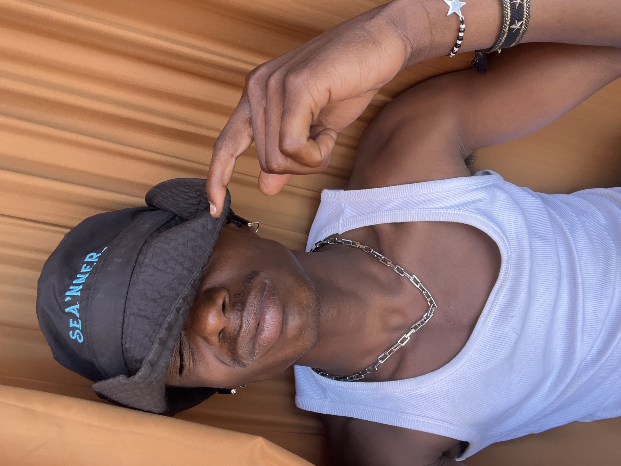A medium close-up shot captures a person lying down, their body oriented diagonally across the frame. The subject has dark skin and is wearing a white, ribbed tank top and a black bucket hat. A silver chain necklace rests on their chest. On their right wrist, two bracelets are visible: one features a silver star charm and black and white beads, while the other is a woven band with gold star accents on a dark background. The person's right hand is raised, with fingers lightly touching the brim of the hat.
The hat has light blue text embroidered on it that reads "SEA'NNER". The person's face is visible, with a light mustache and stubble around their mouth and chin. Their lips are slightly parted. The background is a warm, solid orange-brown color with subtle horizontal stripes or textures, suggesting a soft surface like a hammock or fabric. The lighting is bright and even, indicating daytime. The overall impression is relaxed and casual. There are no direct visual cues to confirm the Barcelona, Spain location context within the image itself.