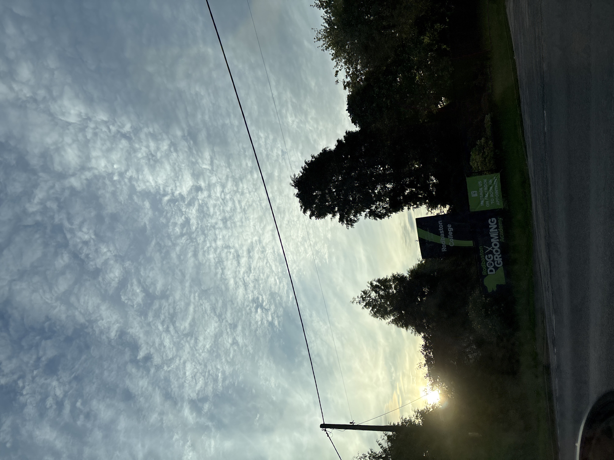 The image shows a street scene in South Staffordshire, United Kingdom, under a cloudy sky. In the foreground, a road is visible, with a slight curve to the right. To the left of the road, there is a silhouette of a telegraph pole with wires extending across the frame. Behind the pole and further back, trees are silhouetted against the sky, with the sun peeking through them on the left side of the image, creating lens flare. In the middle ground, a dark green sign with white lettering is visible, which reads "Rodbaston College" at the top. Below that, another sign in bright green with white lettering says "RODBASTON DOG GROOMING ACADEMY". To the right of the signs, more trees are silhouetted against the sky. The overall impression is of an outdoor, possibly rural, setting at dusk or dawn.