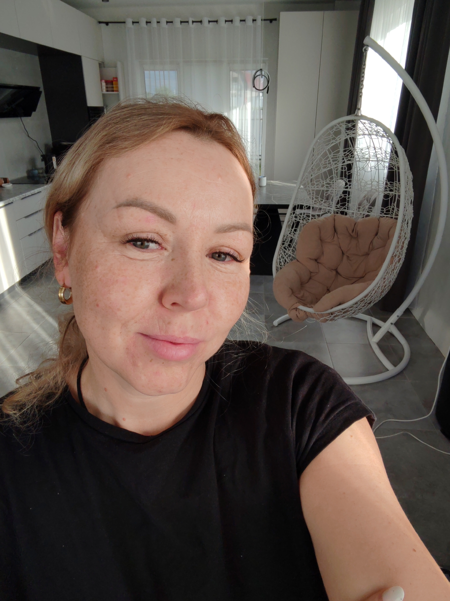 A selfie shows a woman with blonde hair and fair skin. She wears a black T-shirt and gold hoop earrings. The background includes a modern kitchen with white cabinets and a dark countertop. A hanging white chair with a beige cushion is also visible. Based on the metadata, the location is Chaikovsky, Russia.