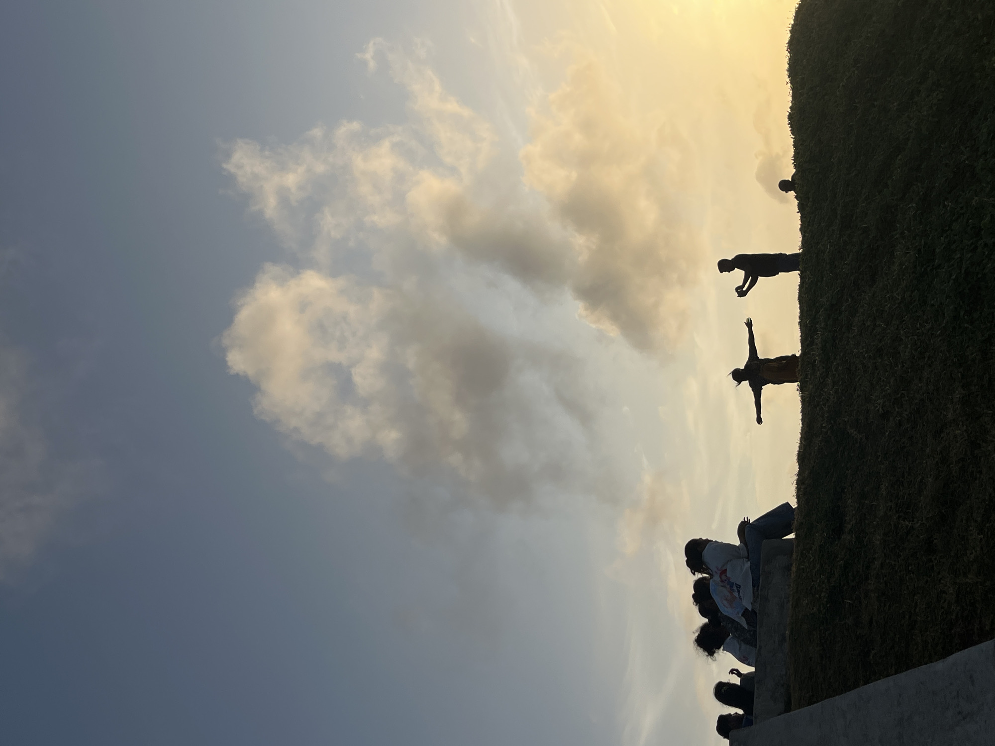 A silhouetted group of people is gathered on a grassy hill under a pale blue sky with scattered, fluffy clouds tinged with yellow from the setting sun. On the left side of the frame, a group of five individuals are seated on a low wall, facing away from the camera. Further to the right, on top of the hill, two figures stand. One figure is facing the camera with arms outstretched, while the other is holding up a phone, possibly taking a picture. Another smaller figure is visible further in the background on the right. The foreground shows a dark, grassy slope with a concrete structure on the far left. The scene appears to be a scenic overlook, potentially in Galle, Sri Lanka, given the warm, tropical lighting and the open sky.