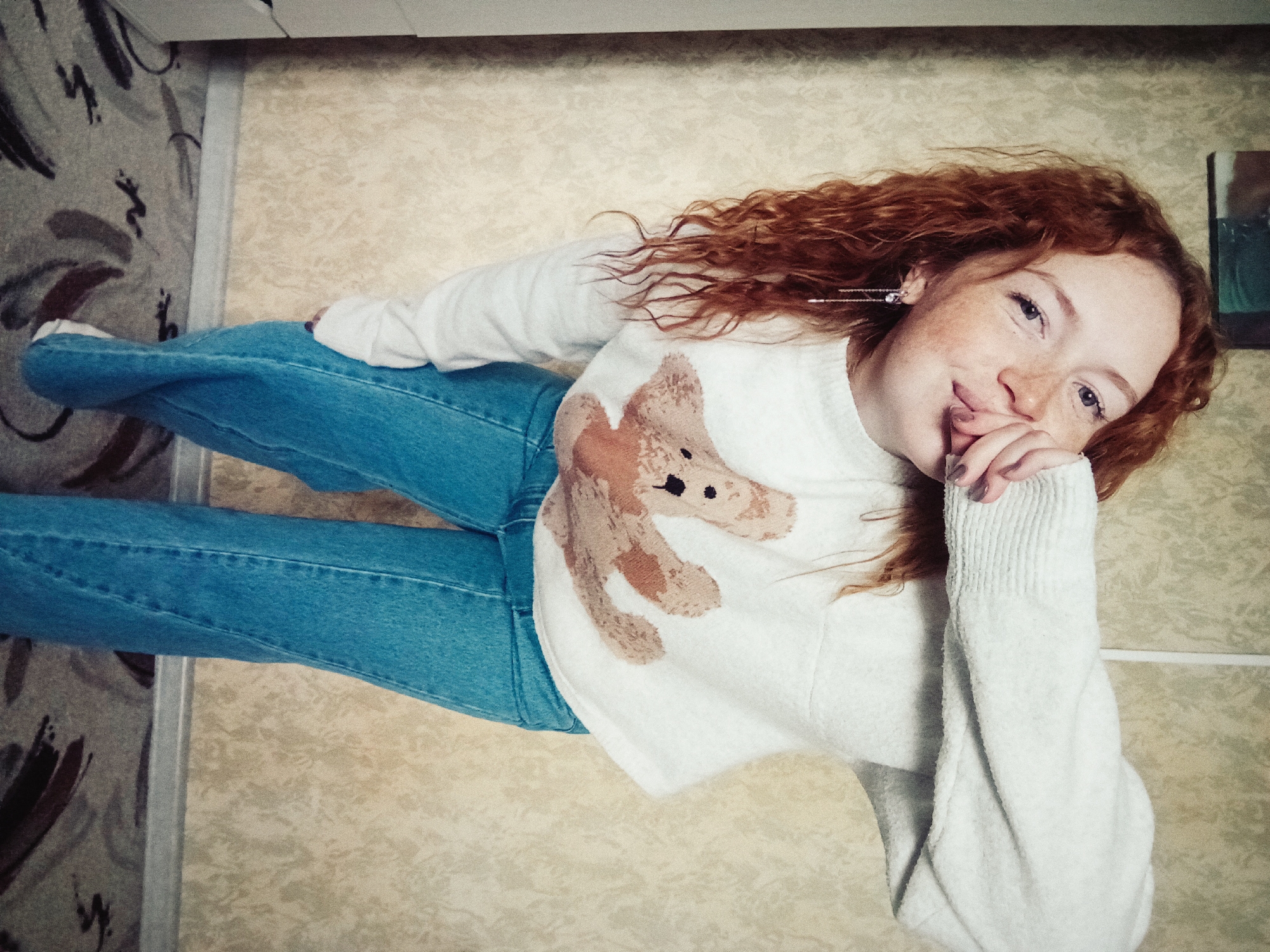 A young woman with curly red hair and freckles is depicted lying down, looking up towards the camera with a soft, engaging smile. She has light-colored eyes and her right hand, with light-colored nail polish, is gently placed near her mouth. She is wearing a white long-sleeved sweater adorned with a large, light brown teddy bear design on the front, paired with light blue denim jeans. Her left ear features a long, dangling earring.
The setting is indoors, with the perspective looking down upon the woman. The background is divided into two main areas. To the left, there is a carpet or rug with a greyish base and prominent abstract patterns in dark brown and black. To the right, there is a lighter, beige-toned surface with a textured, subtle pattern, likely wallpaper or flooring. A narrow, light-colored strip separates these two background elements. A small, dark-framed object, possibly a picture or mirror, is partially visible in the upper right corner of the image. The lighting is soft and consistent, suggesting an indoor, possibly well-lit, environment. There is no visible text in the image. The overall impression is one of comfort and warmth, with the woman conveying a friendly and relaxed demeanor.