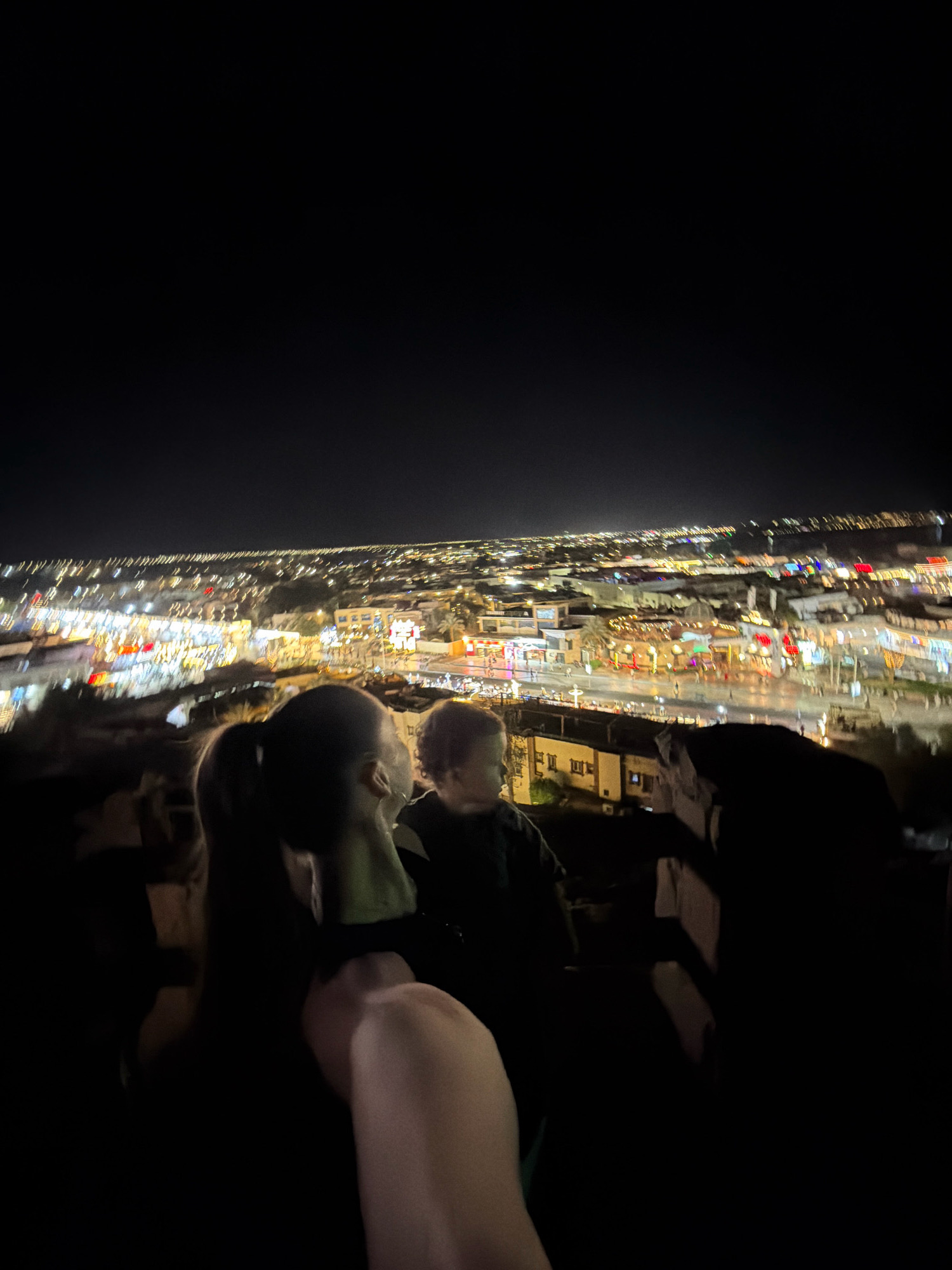 An aerial nighttime shot captures a panoramic view of Sharm El Sheikh, Egypt, with its cityscape illuminated by a multitude of lights. The foreground is dominated by the silhouettes of several individuals, obscuring much of their features. Their backs are to the camera, as they appear to be observing the city below. The middle ground reveals a densely packed urban area with buildings of varying sizes and architectural styles, interspersed with palm trees. The city is ablaze with light, suggesting a vibrant commercial or residential district. Numerous streetlights, illuminated signs, and building lights create a dazzling spectacle. The overall impression is one of a lively and energetic city at night.