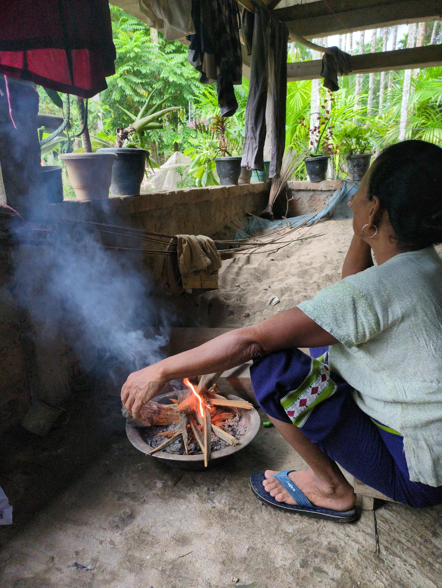 A woman sits on the sandy floor of an open-air structure, tending to a small fire in a shallow metal basin. She wears a light green patterned shirt and dark blue pants with a decorative band on the leg. Her right arm is extended, and her hand is near the fire, adjusting the wood. A plume of smoke rises from the fire. In the background, various potted plants are visible, and beyond them, lush green vegetation and palm trees suggest a tropical environment. The structure appears rustic, with a wooden roof and exposed beams, and clothes are hung on a line overhead. The scene is set in Borchora, India.