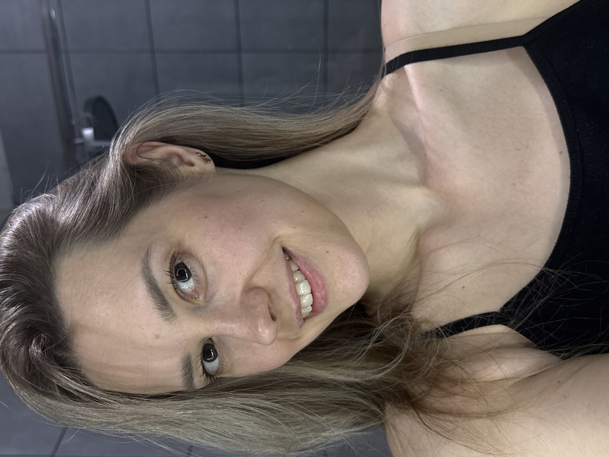 A close-up portrait shows a young woman with a joyful expression, looking directly at the viewer. Her head is tilted, giving the image a dynamic, candid feel, possibly a selfie. She has long, light brown hair, which frames her face and flows over her shoulders and chest. Her eyes are dark, and she is smiling broadly, revealing white teeth. Her fair skin shows a few subtle freckles or moles.
She is wearing a black tank top or sports bra with thin straps, one of which is visible over her right shoulder and collarbone. A small, decorative earring, possibly gold-toned and star-shaped, is visible in her right ear.
The background is a dimly lit indoor setting, appearing to be a dark, possibly tiled wall, with a faint, blurry reflection in the upper left corner that might suggest a mirror or a window. The focus is entirely on the woman, with the background receding into soft blur. The overall impression is one of happiness and casual intimacy. No text is visible in the image. Given the location context of Chaikovsky, Russia, the subject is presumably located there.