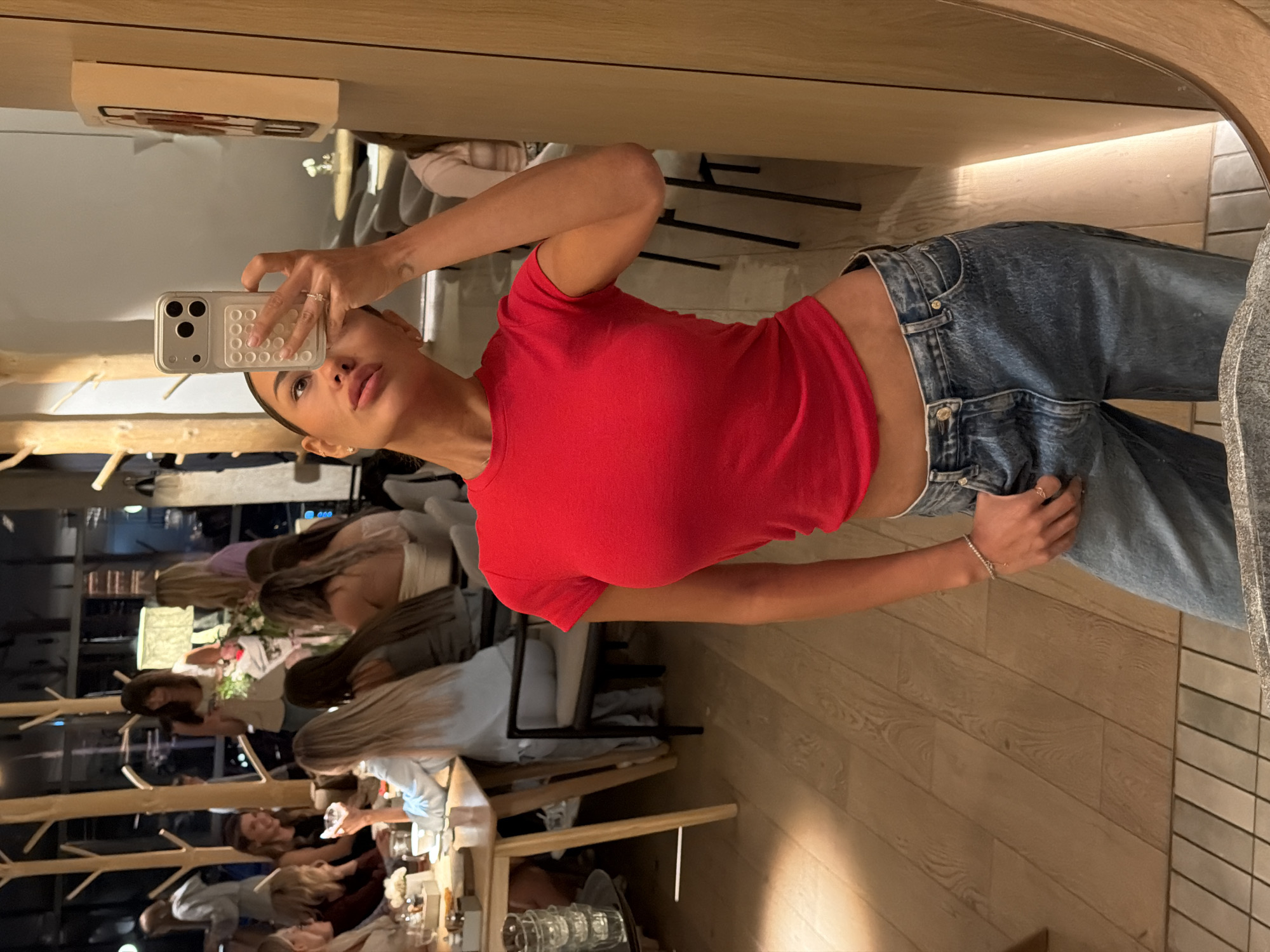 The image depicts a woman in a red top and blue jeans taking a mirror selfie in what appears to be a restaurant or a similar indoor public setting. The woman is positioned in the foreground, looking into a large mirror on the right side of the frame. She is holding a smartphone in her right hand, capturing the reflection. Behind her, through the mirror's reflection and also visible directly in the background, other individuals are seated at tables, suggesting a social gathering. The environment features wooden tables, chairs, and decorative elements that resemble trees, creating a modern and possibly botanical-themed interior. The lighting suggests it is evening or nighttime. The image does not contain any specific visual cues that would allow for pinpointing its location to Krasnoyarsk, Russia.