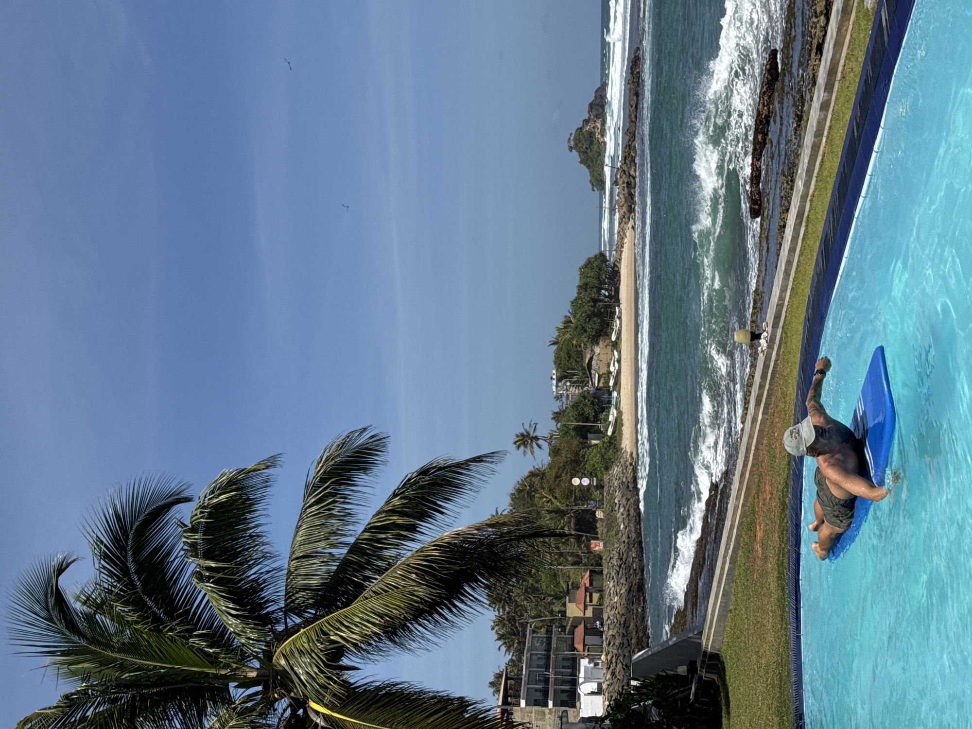 A man wearing a baseball cap and olive green shorts is lying face down on a blue inflatable mat in a bright blue swimming pool. He is positioned in the lower left quadrant of the frame, with his arms extended forward and his head turned to his left. The pool is surrounded by a grassy area and a concrete ledge. In the background, there is a turbulent ocean with waves breaking on a rocky shore. A beach with sparse vegetation and some buildings is visible further down the coast. A small, rocky island with a few trees is situated in the ocean. Palm trees are prominent in the scene, with one large palm tree arching over the left side of the frame. The sky is clear and blue with a few distant birds. The location is Goviyapana, Sri Lanka.