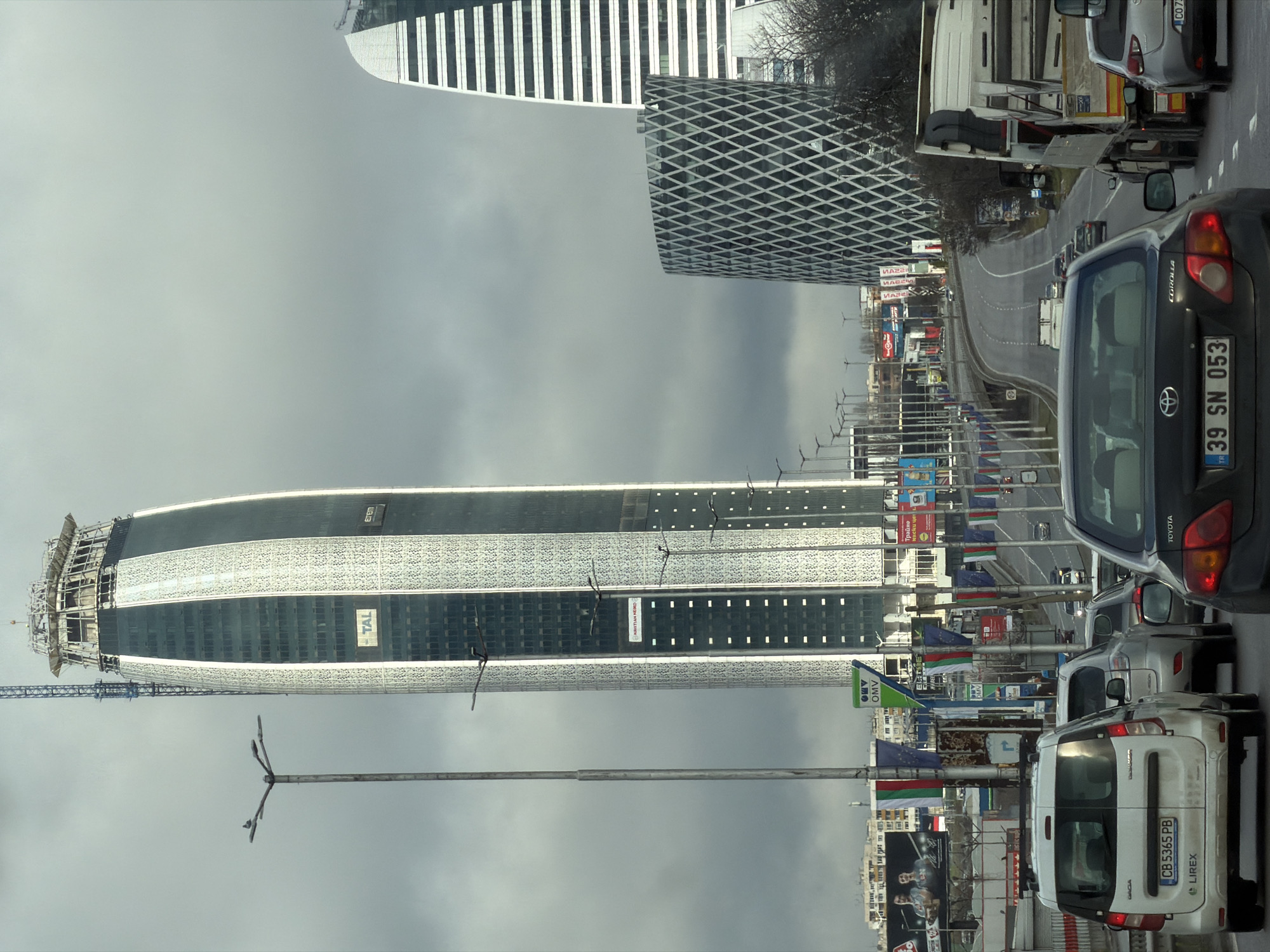 The image captures a scene in Sofia, Bulgaria, dominated by the presence of several prominent skyscrapers against a cloudy sky. Visible are the Capital Fort, Sky Fort, and another building featuring a distinctive diamond-patterned facade. In the foreground, a busy street is filled with traffic, including a Toyota Corolla with the license plate "39 SN 053" and a white Dacia van with the license plate "CB 5365 PB." Flags and advertisements line the street, and construction is evident on one of the skyscrapers.