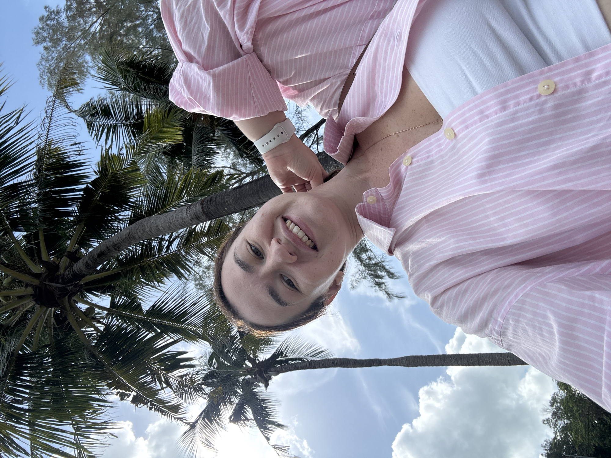 A woman with brown hair and fair skin is taking a selfie outdoors under palm trees and a blue sky with scattered clouds. She is smiling and wearing a pink and white striped button-down shirt over a white tube top, and a white smartwatch on her left wrist. She has a thin gold chain necklace visible. The image was taken in Pa Khlok, Thailand, with palm trees and lush foliage framing the scene.