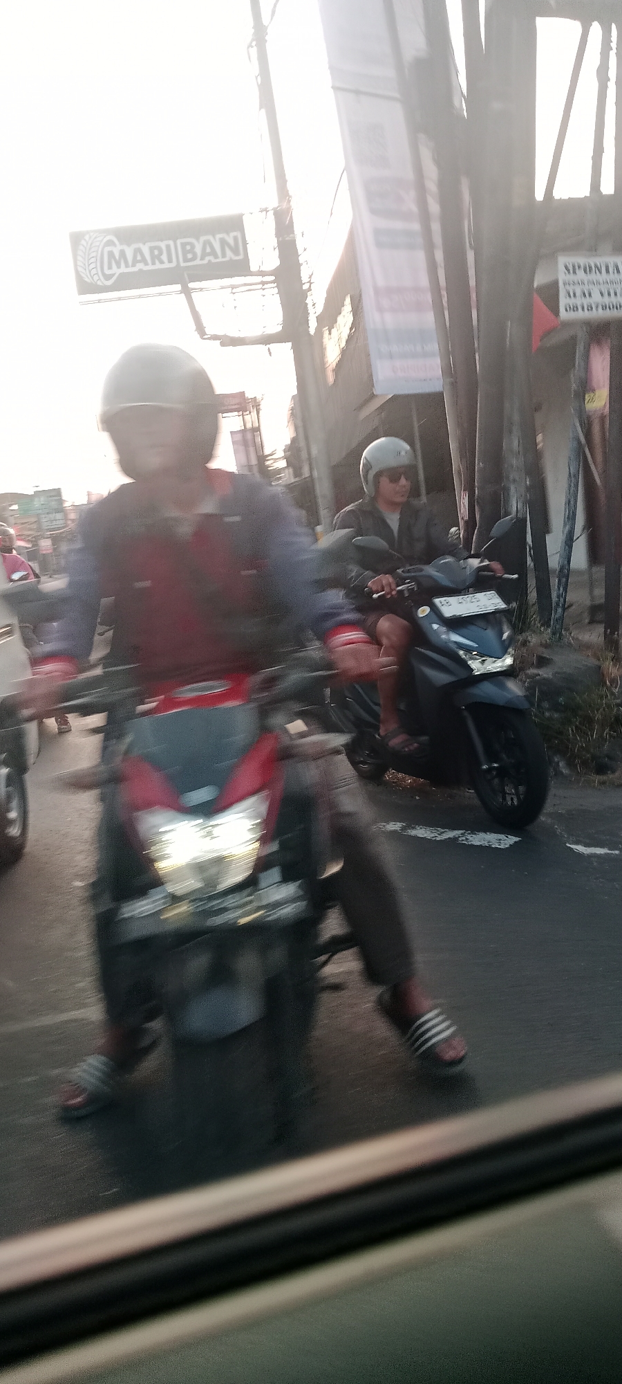 In Banyuraden, Indonesia, a medium shot captures a street scene with motion blur, indicating movement. Two individuals on motorcycles are the primary subjects. In the foreground, a person wearing a dark helmet and a red and blue jacket is riding a red motorcycle. Their hands are on the handlebars, and their legs are visible, wearing dark pants and striped sandals. The motorcycle's headlight is illuminated. Behind this motorcycle, another person wearing a light gray helmet and sunglasses rides a dark gray motorcycle. This person is dressed in a dark jacket and shorts, with their legs also visible and wearing sandals. They are holding the handlebars. The background shows a street with signage, including a banner that reads "MARI BAN" with a tire graphic, and another sign with Indonesian text. Sunlight streams in from the right, casting lens flares and creating a bright, hazy atmosphere. The road surface is visible, with some vegetation on the right side. The overall impression is of a busy street with traffic in motion during daylight.