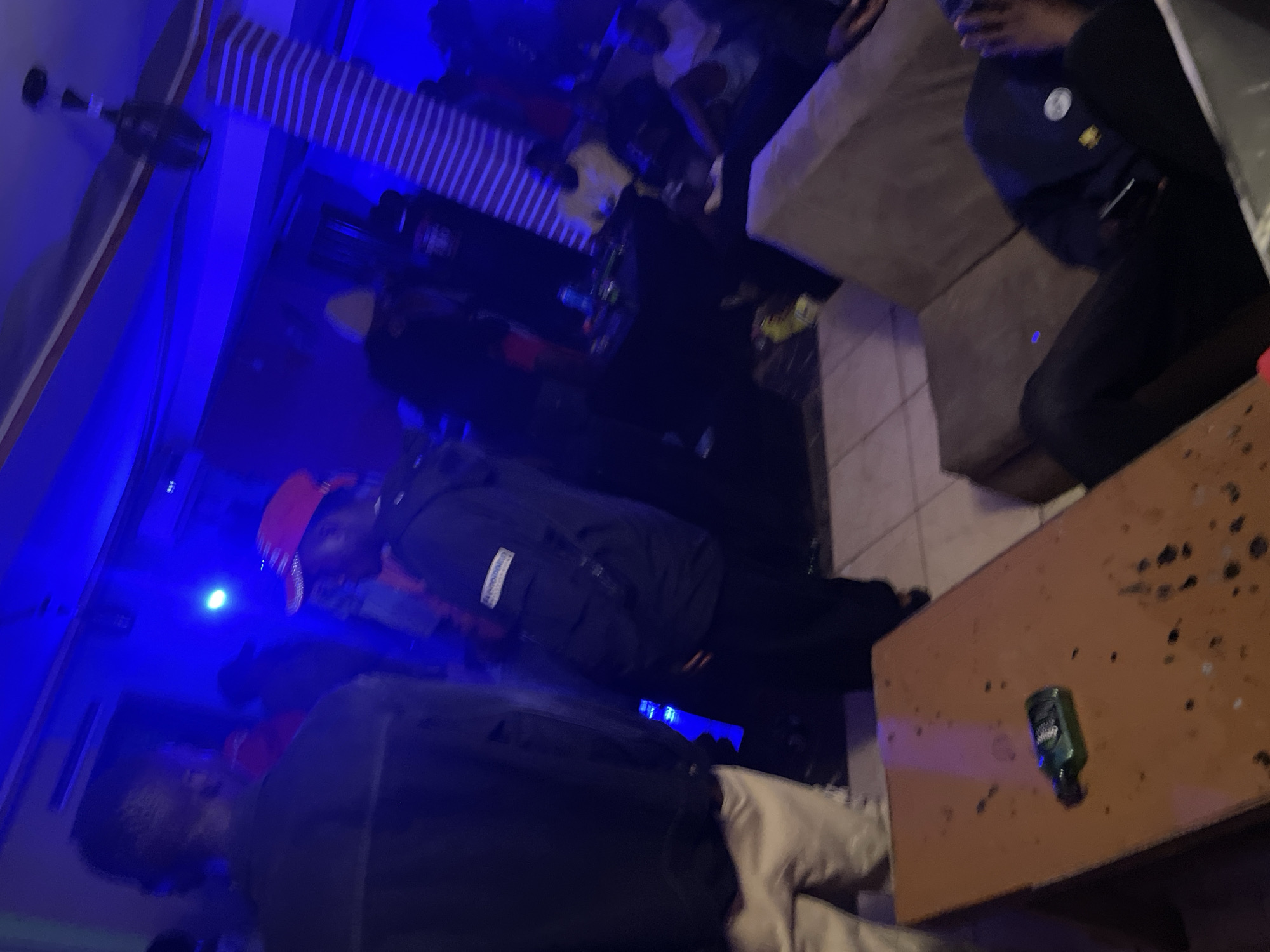 The image depicts a dimly lit indoor space, likely a club or lounge, illuminated by vibrant blue lighting. Several individuals are present, some standing and others seated or reclining. In the foreground, a table holds a small green bottle. The overall atmosphere appears to be one of social gathering, possibly a party or casual hangout. The location is specified as Durimi, Nigeria.