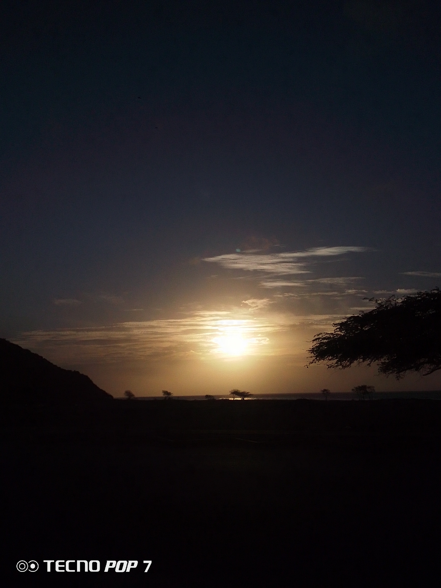 The image depicts a landscape at dusk or dawn, likely near Pedro González, Venezuela. The sky is a gradient from dark blue to light orange where the sun is visible, obscured by thin clouds. Silhouetted against the sky are trees, a hill to the left, and what appears to be a body of water on the horizon. The foreground is dark. The text "TECNO POP 7" is in the lower left corner.