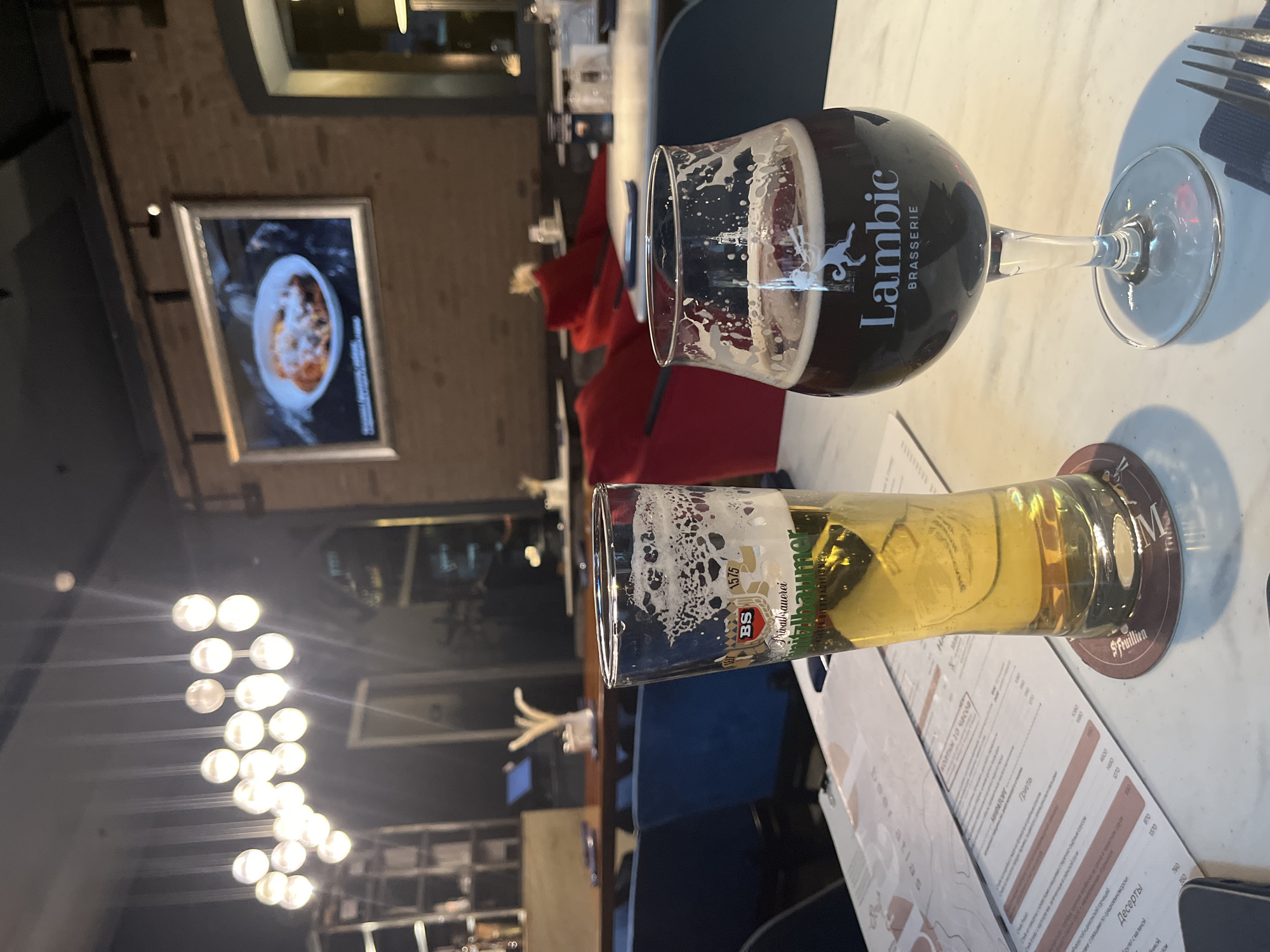 The image shows a table setting at a restaurant in Moscow, Russia. On the table are two glasses of beer: one a dark Lambic and the other a light-colored beer labeled "Privatbrauerei" with the "BS" logo. A menu is visible with prices in rubles, including dessert options. In the background, there is an artwork depicting food, a lighting fixture, and furniture indicating a restaurant interior. The photograph is slightly out of focus in the background. There is a fork on the table, indicating a meal setting.