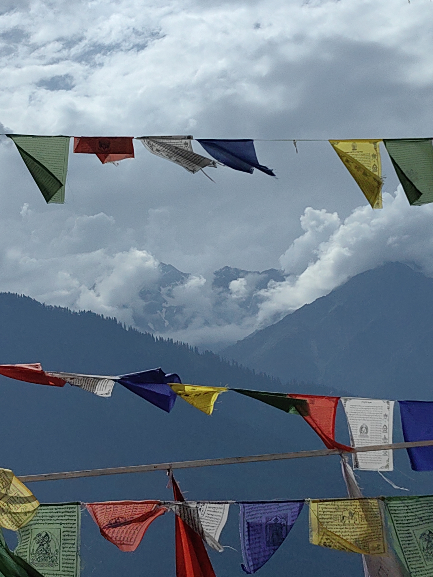 In Siumsa, India, prayer flags flutter against a backdrop of cloudy, mountainous terrain. The image is divided into two horizontal sections by the flags. In the upper section, green, red, blue, and yellow prayer flags are strung across a wire, partially obscuring a sky filled with white clouds and distant, shadowed mountains. The lower section features another row of prayer flags, predominantly in red, blue, yellow, and green, with white flags bearing text and images interspersed. These flags are also strung across a wire and are in the foreground, with the dark blue-gray of the mountains and a dark forest visible behind them. The overall scene suggests a high-altitude location with a mix of natural beauty and cultural elements.