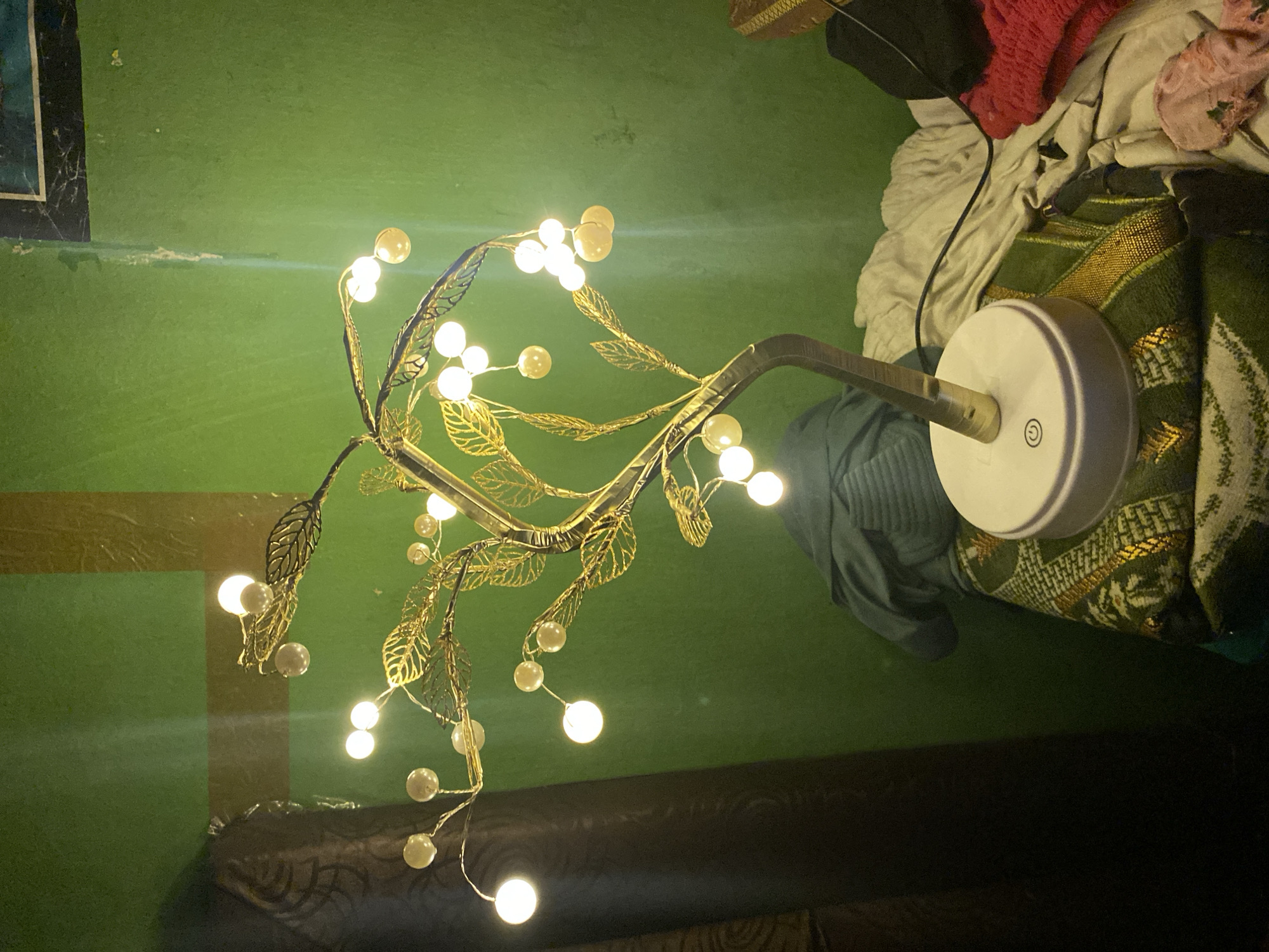 The image captures a brightly lit decorative lamp in an indoor setting, likely in Tanjongkapai, Indonesia. The lamp features a metallic, tree-like structure with numerous delicate gold-colored leaves and small, spherical lights, some glowing intensely white, others appearing as opaque pearls. The lamp's stem connects to a circular white base which has a power symbol (a circle with a vertical line) printed on it.
The dominant background is a textured, solid green surface, possibly a wall or floor, showing some minor scuffs and imperfections. To the right of the lamp, a collection of textiles and clothing is visible, including a dark green fabric with intricate gold patterns, a crumpled off-white cloth, and a section of bright red clothing. A black power cord snakes through these fabrics. In the upper left corner, against the green background, a portion of a dark-framed object or picture is visible.
The bright illumination from the lamp suggests it is operating in a dimly lit environment, possibly at night. No people are present, and no specific activity is taking place beyond the lamp providing light to the immediate area. The scene depicts a segment of an interior space, characterized by the lamp's unique design and the surrounding household items.