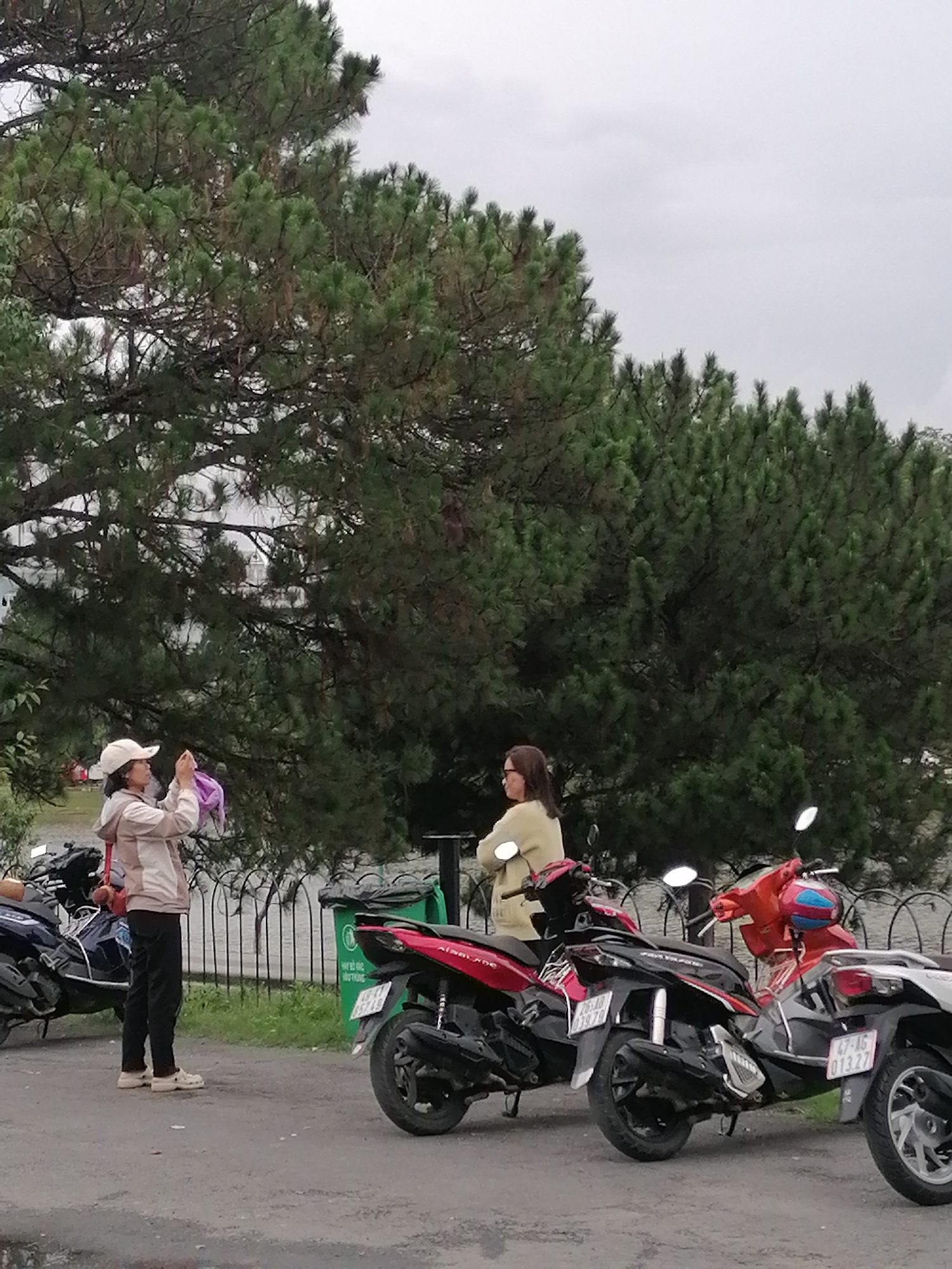The image shows a street scene in Da Lat, Vietnam. Two women are standing near several parked scooters. One woman, wearing a tan jacket and a white cap, is holding a purple item and appears to be taking a photograph. The other woman, wearing a light yellow sweater, has her arms crossed. The background includes a large tree, a fence, a body of water, and an overcast sky. A green trash can is visible near the fence. The scooters are various colors including red, black, and white. License plates are visible on some of the scooters.
