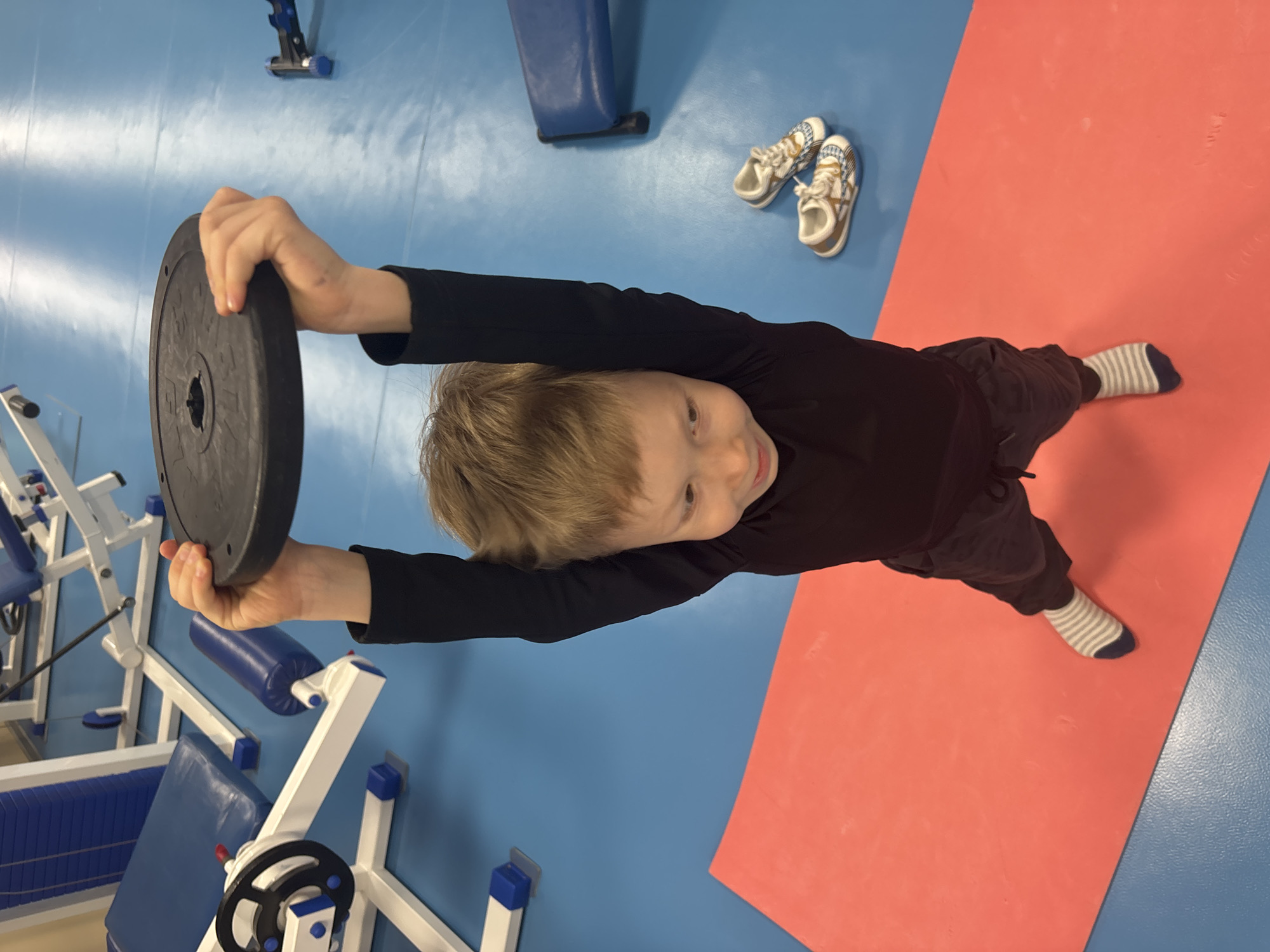 A young boy in a black long-sleeved shirt and dark pants, wearing striped socks, lies on a red mat in a gym-like environment. He holds a black weight plate with both hands, extending his arms straight above him. The floor is blue, and gym equipment is visible in the background, including weight machines with blue padding and metal frames. A pair of children's shoes are present near the corner of the mat. The location is the city of Chaikovsky, Russia.