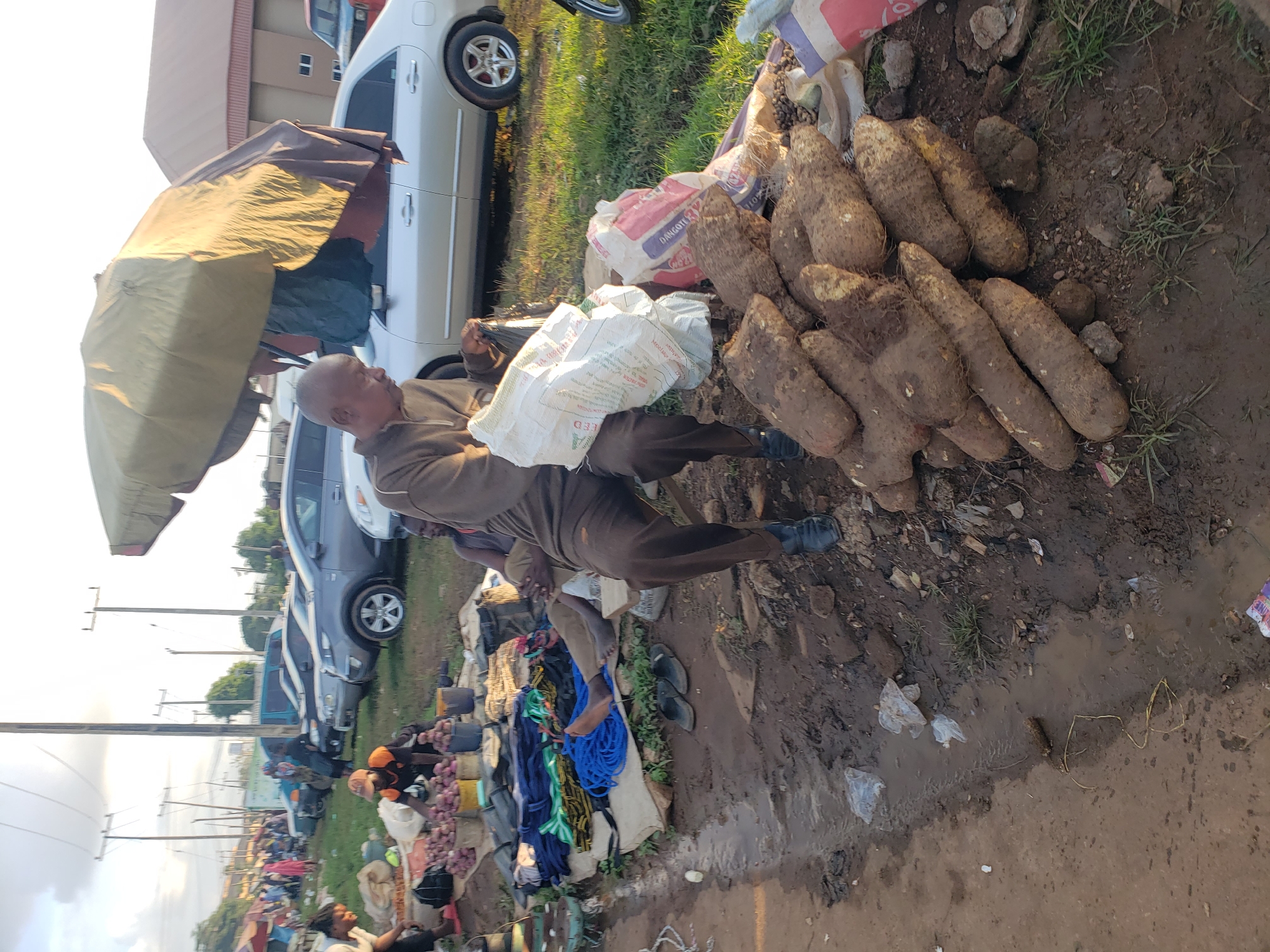 The image depicts an outdoor market scene in Kakuri Gwari, Nigeria. In the foreground, a pile of yams is displayed on the ground. To the left of the yams, a man is sitting on a low wooden stool, holding a white bag. Behind him, another man is seated on a sack, partially obscured. Further back, various goods including ropes and what appear to be onions in buckets are visible on the ground. In the background, several cars are parked, and market stalls with canopies are set up. The ground is muddy and uneven, with some puddles of water. Power lines and poles are prominent in the sky, indicating an urban or semi-urban setting. The overall impression is of a busy marketplace with a variety of goods and people.