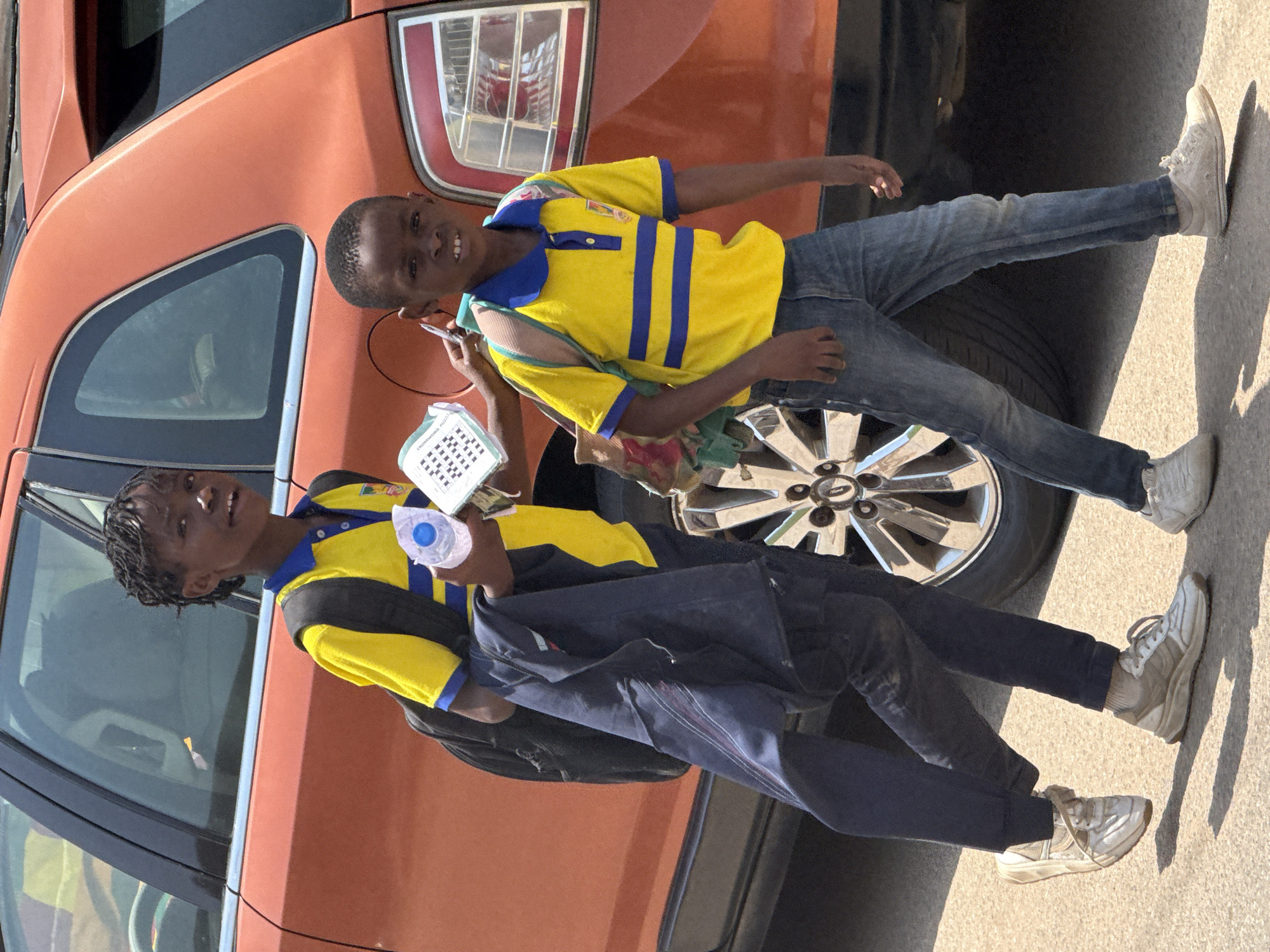 Two young children, likely students, walk along a street in Abuja, Nigeria, with an orange car parked behind them. The child on the left, taller with braided hair, wears a yellow polo shirt with blue stripes, dark pants, and a black backpack. They are carrying a dark blue jacket and a water bottle. The child on the right, shorter, wears a similar yellow and blue striped polo shirt, blue jeans, and a backpack. Both children are wearing white sneakers. The car, a hatchback, is a vibrant orange color with a visible silver wheel. The ground appears to be asphalt, indicating an urban setting. The lighting suggests it is daytime, with a clear sky.