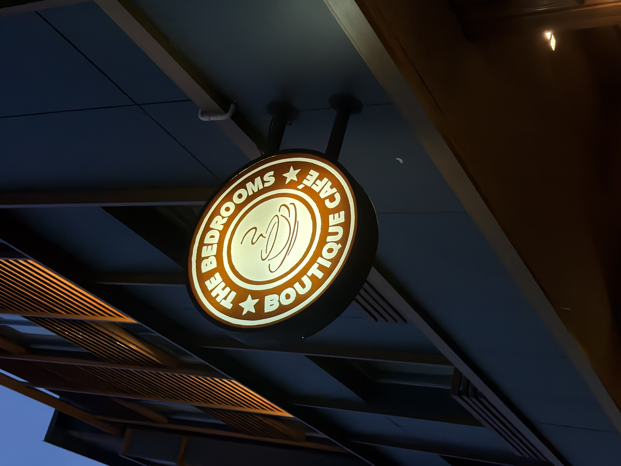 A circular, illuminated sign for "THE BEDROOMS BOUTIQUE CAFÉ" is mounted on the exterior of a modern building at night. The sign features a stylized coffee cup and steam within a starburst motif, surrounded by text. The building's facade is dark, with vertical wooden slats illuminated by warm orange light on the upper levels, and dark panels below. A crescent moon is visible in the night sky. The image appears to have been taken at an upward angle, suggesting the building is multi-story. The context provided indicates this scene is located in Bangkok, Thailand.