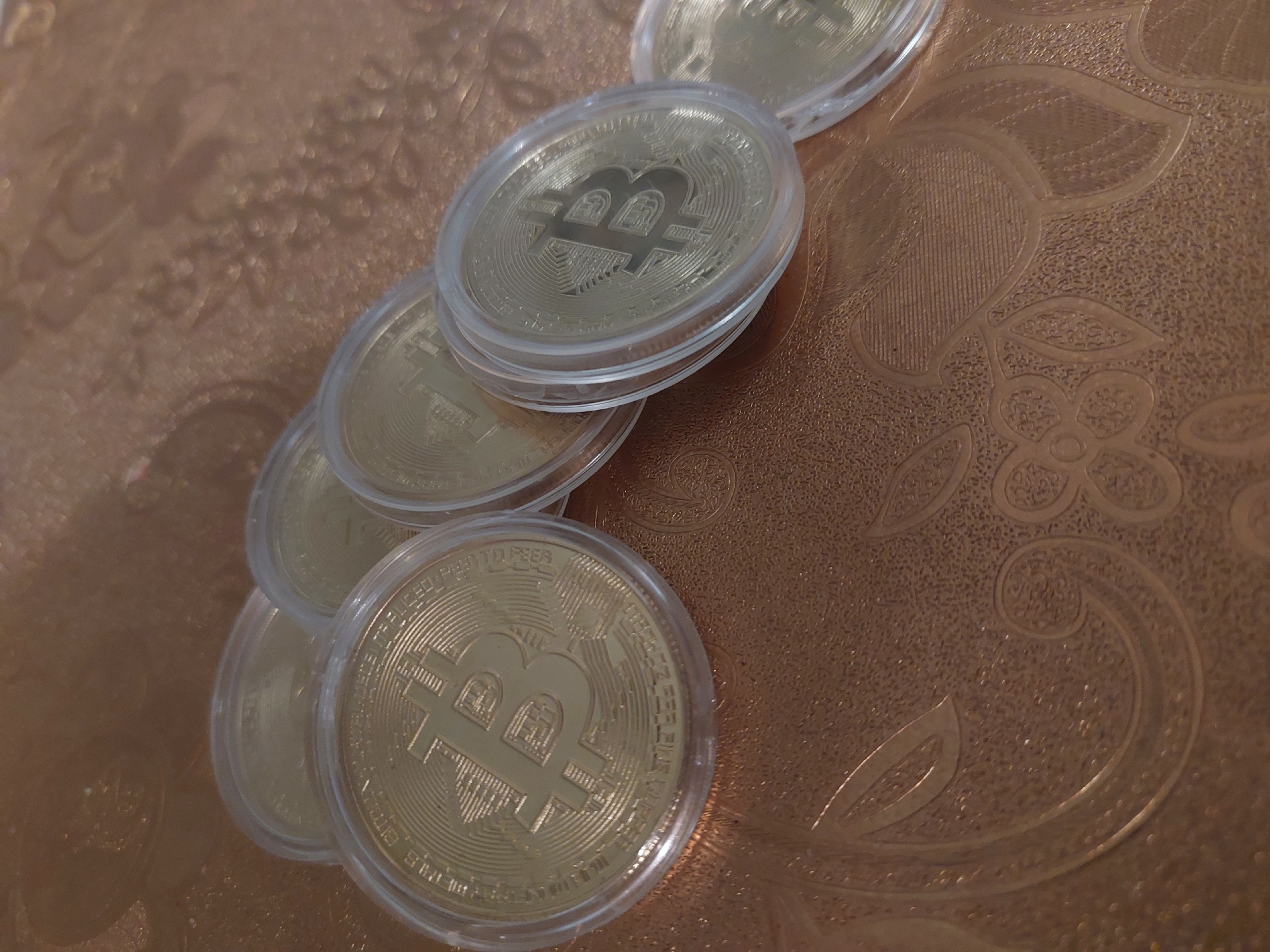 The image shows several physical Bitcoin coins in clear plastic protective cases, arranged on a patterned, reflective, copper-colored surface. The pattern on the surface appears to be floral. The coins are mostly gold in color and display the Bitcoin symbol. The image does not provide any information to indicate a location in Tsarevo, Bulgaria.