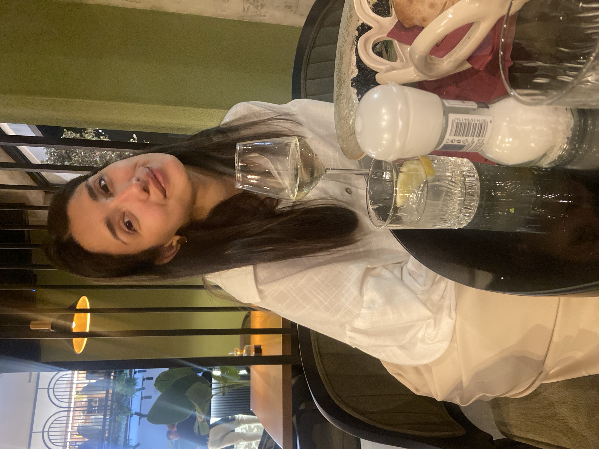 The image, rotated counter-clockwise, depicts a woman seated at a table in what appears to be an evening restaurant or cafe setting in Sutomore, Montenegro. She has long, dark hair and is wearing a light-colored, possibly white, collared shirt. Her face is directed towards the camera with a neutral to slightly pleasant expression.
The table in front of her holds several items: a wine glass containing a clear liquid, another glass with a lemon slice and liquid, and a white salt shaker. The salt shaker features a barcode with the numbers "9 001414 041147" and partial text that reads "JODOSO". Also visible on the table is a white, decorative basket containing bread or similar food, resting on a dark red napkin.
The background suggests a semi-outdoor or well-lit indoor space. Behind the woman, there's a dark, perhaps green, wall and a view beyond metal railings. This view shows artificial lighting, distant structures, and some foliage, indicating it is nighttime or dusk. A yellow-orange light fixture can be seen in the background. The overall ambiance is relaxed and casual, likely during a meal or social gathering.